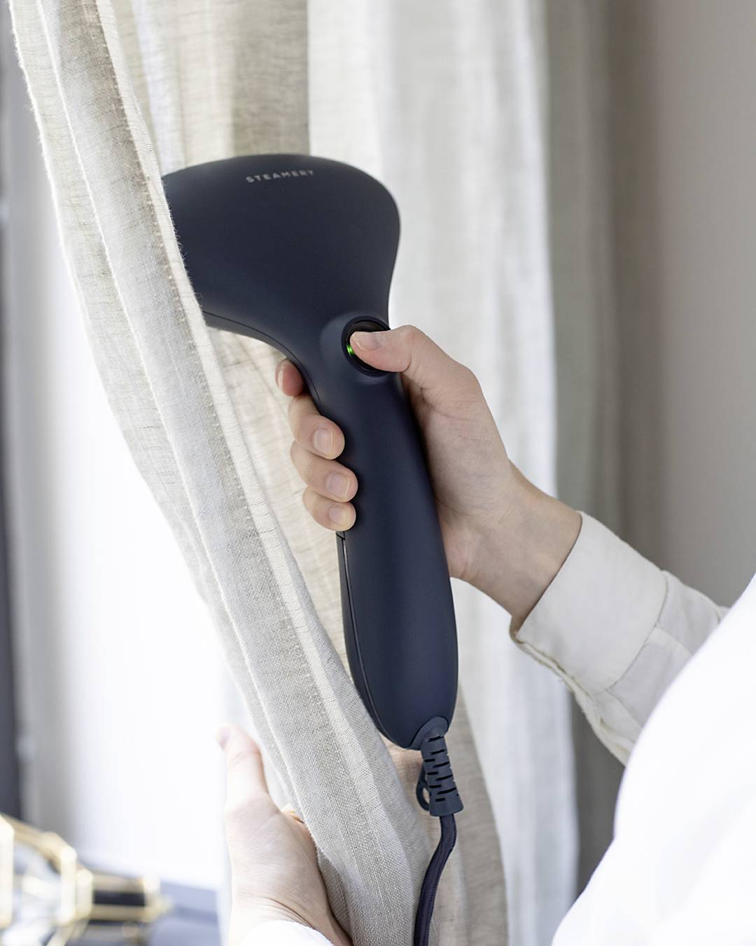 A person is operating a portable steam cleaner and using it to smooth out a beige curtain by a window.