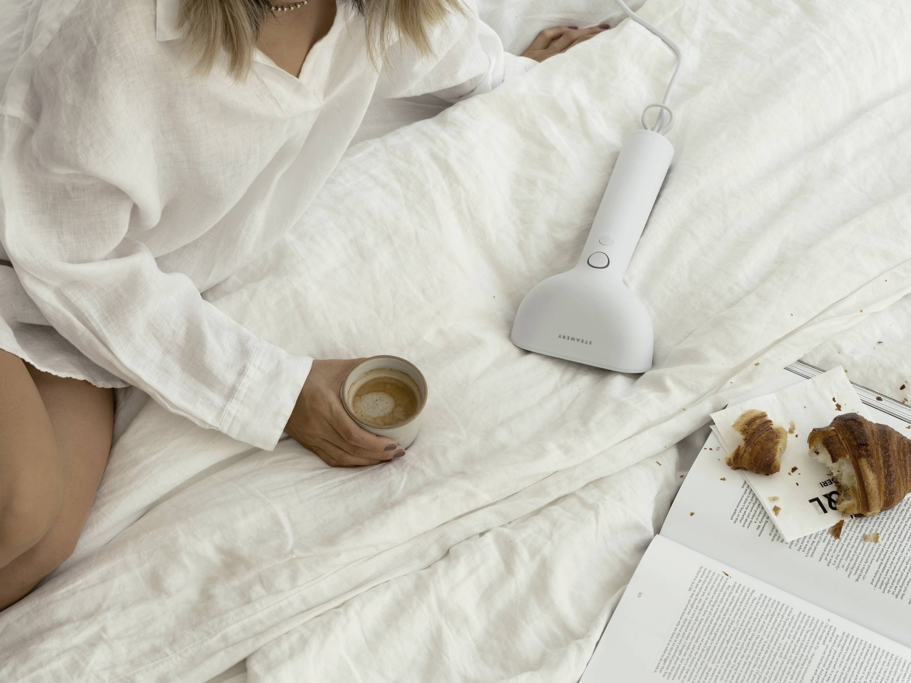 A person in a white dressing gown sitting on a bed, holding a cup of coffee. Next to them is a steam iron, croissants, and an open magazine.