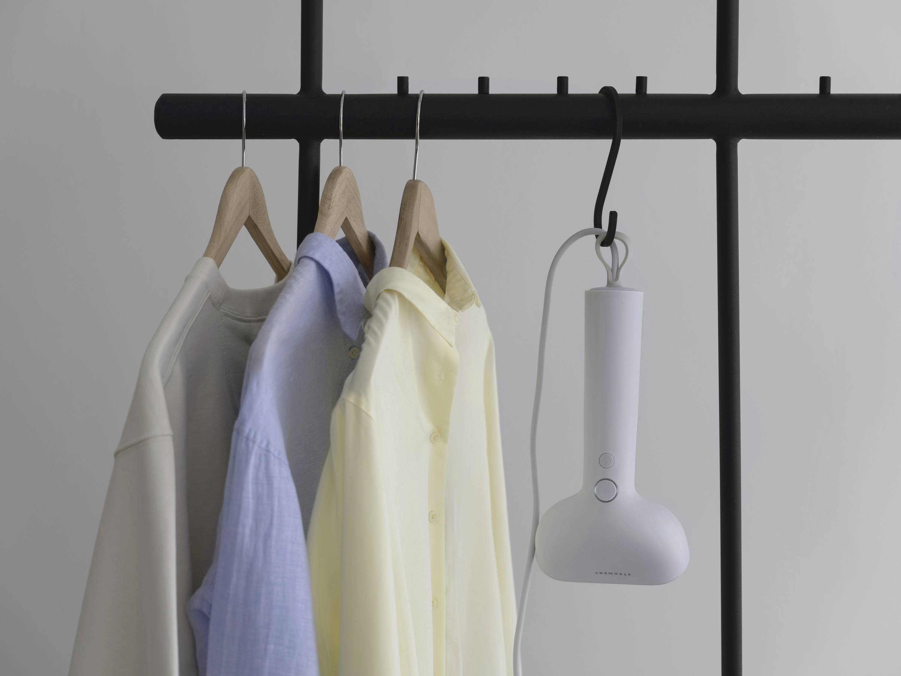 A steam iron is hanging on a clothes rack, beside three shirts in white, blue, and yellow, which are hanging on wooden coat hangers.
