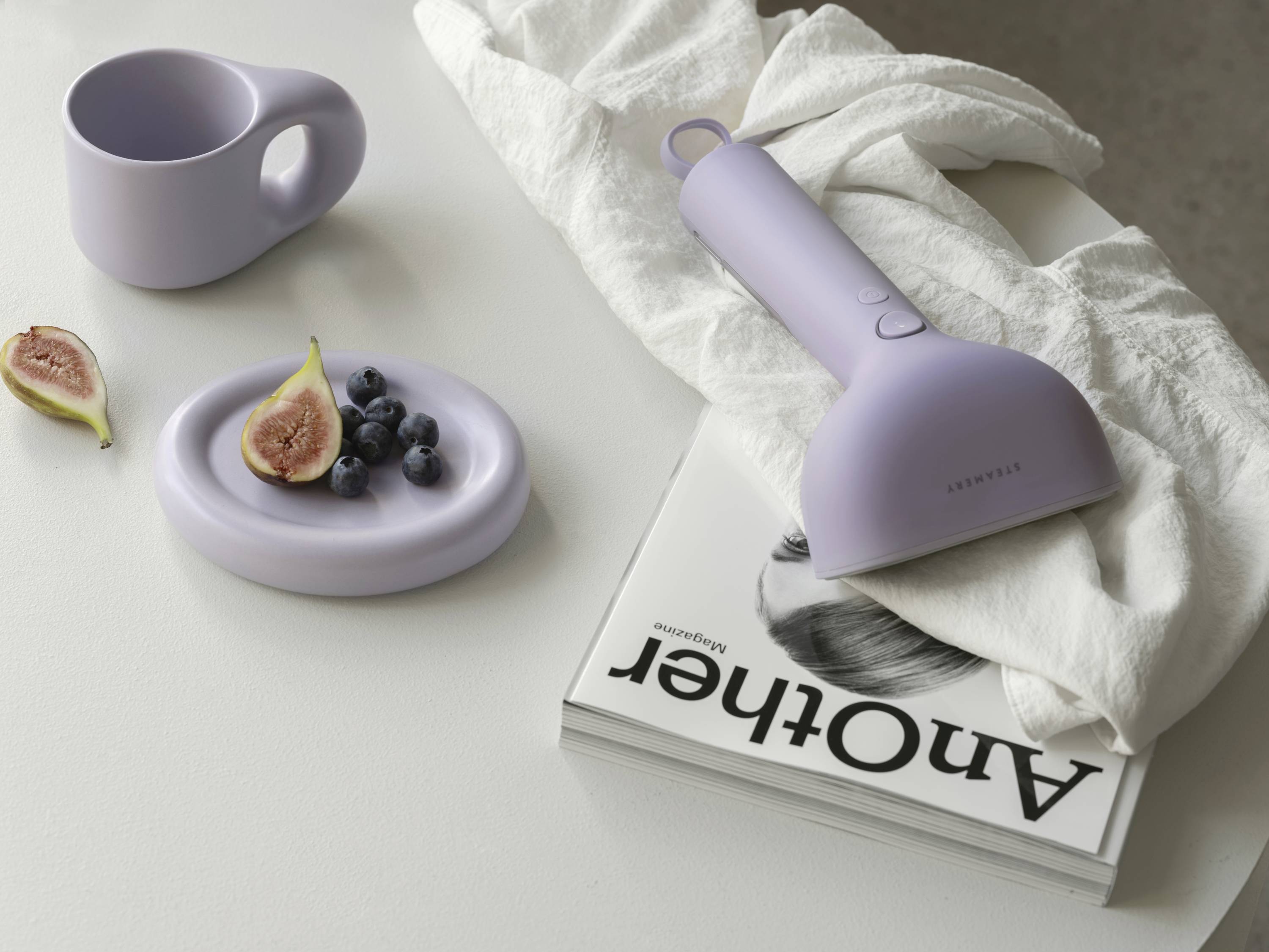 Lila cup, plate with figs and blueberries, handheld vacuum cleaner, 'Another' magazine, white tablecloth; minimalist arrangement.