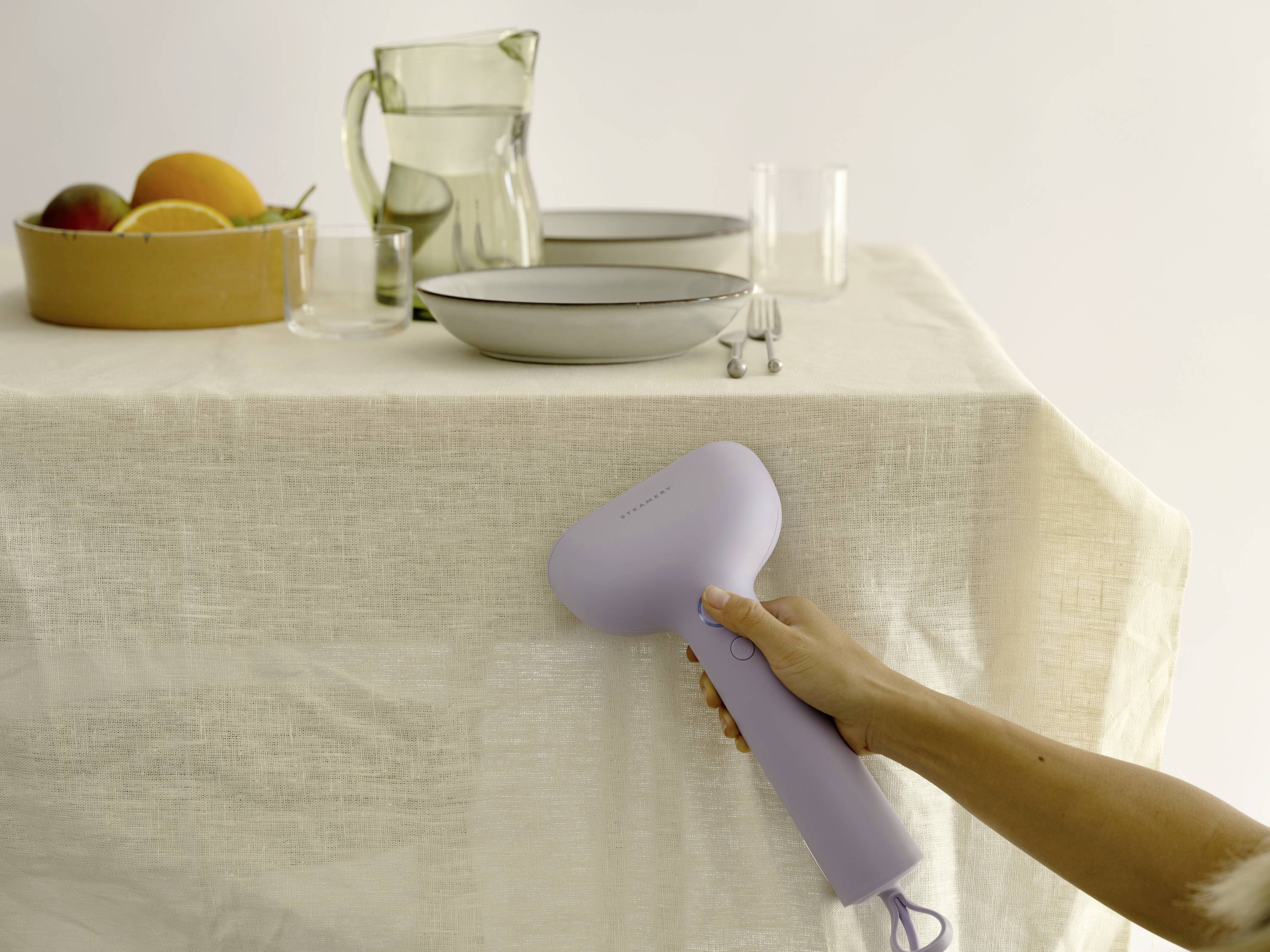 A hand holds a purple clothing steamer in front of a table laid with bowls of fruit, a jug, plates, and glasses.