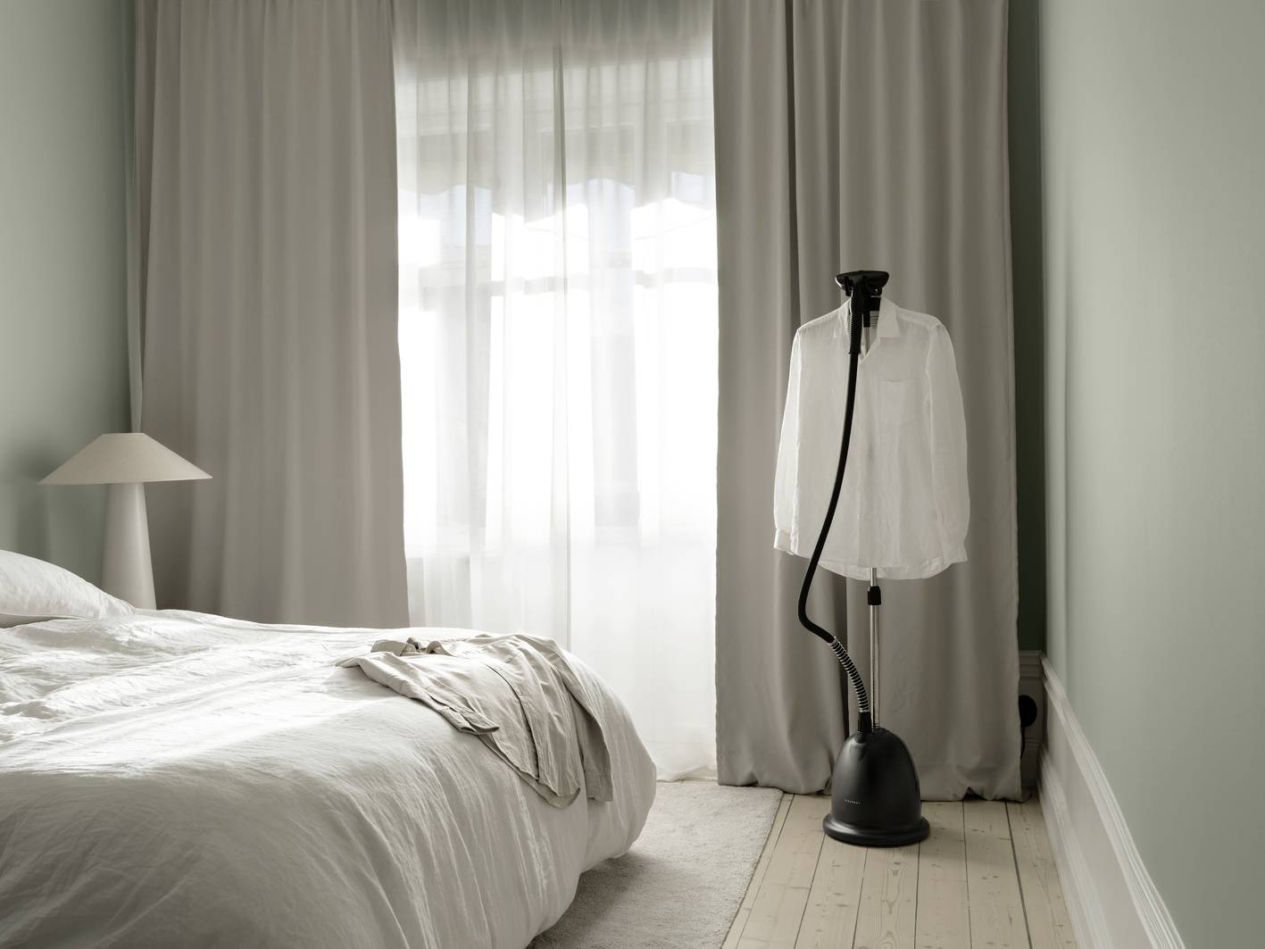 A simply furnished bedroom with a bed and a clothing steamer, on which a white shirt hangs, in front of a window.