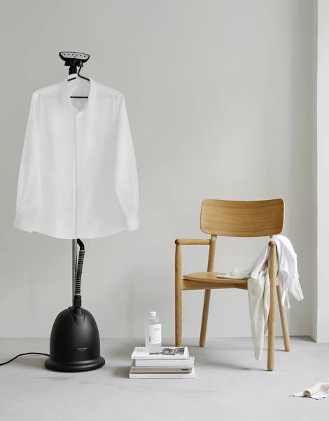 A shirt on a steam iron next to a wooden chair with a towel. Books and a cleaning product are on the left. Minimalist room.