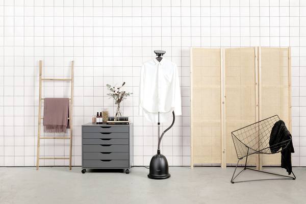 A room with a clothes stand and a white shirt. To the left, there is a ladder with a towel and a chest of drawers, and to the right, a black chair.