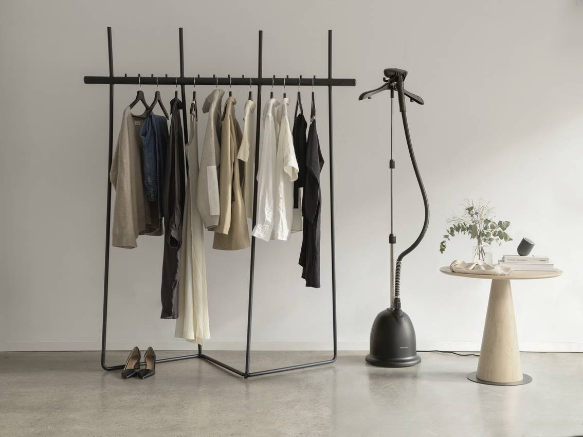 A minimalist wardrobe stands on a concrete floor, with various coloured shirts. Next to it, a steam iron and a round table with a vase.