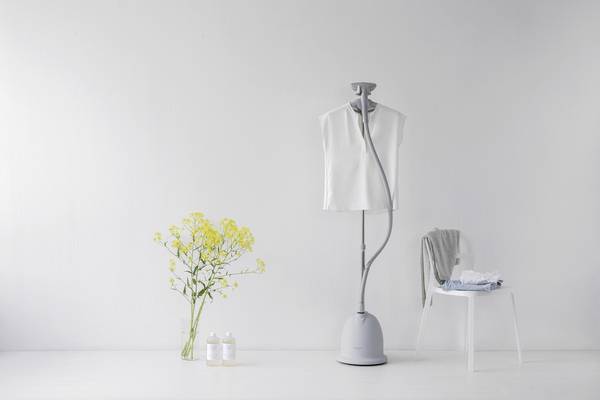 A white sleeveless top hangs on a steaming ironing system. Next to it stands a table with folded clothing and a bouquet of yellow flowers.