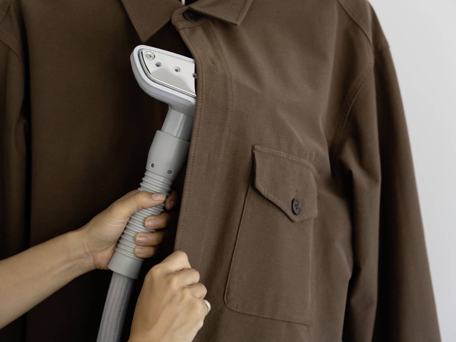 A person is steaming a brown shirt with a handheld steamer. The steam iron is carefully guided over the front of the shirt.