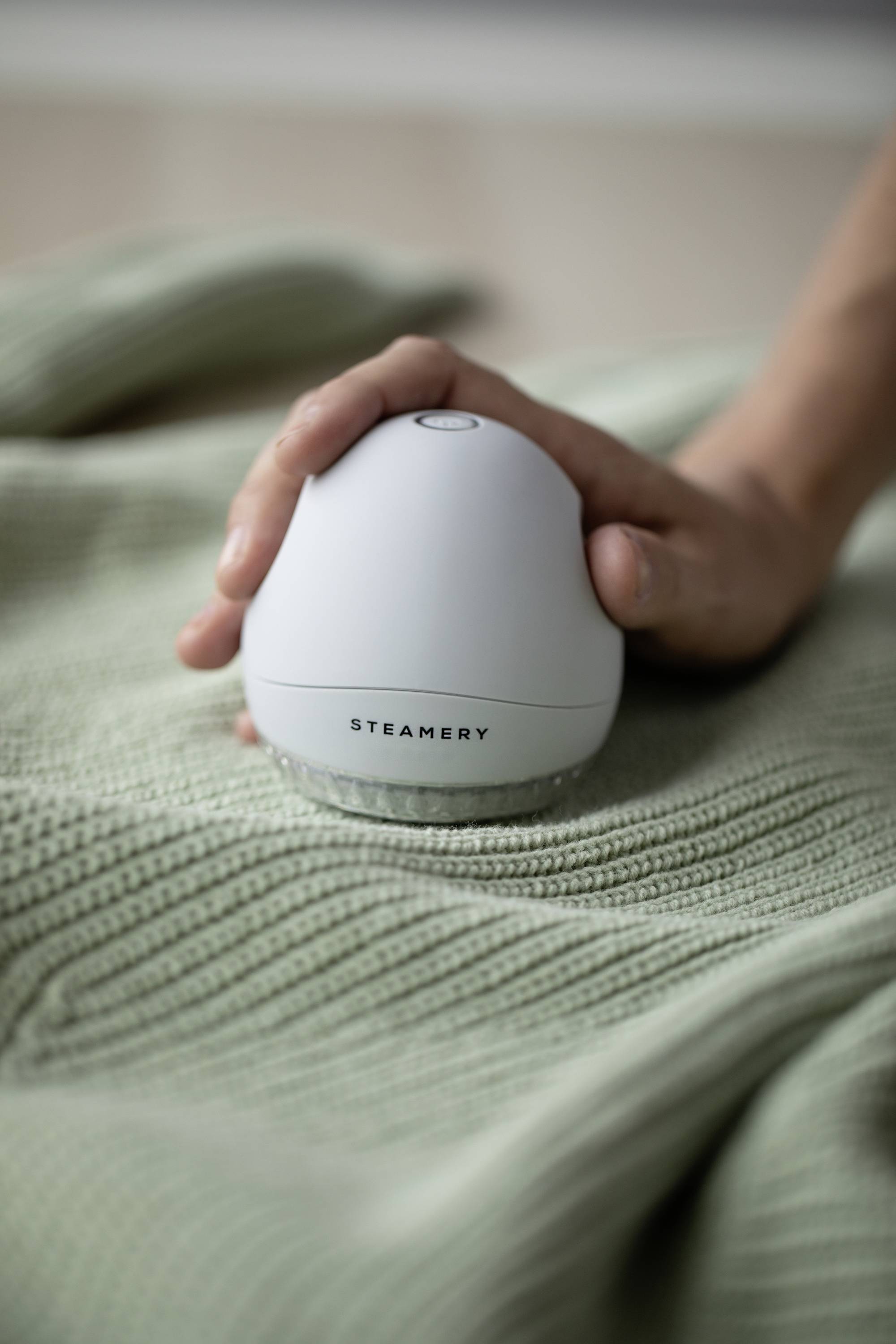 A hand is holding a white fabric shaver over a green knitted jumper. The device bears the text 'STEAMERY'.