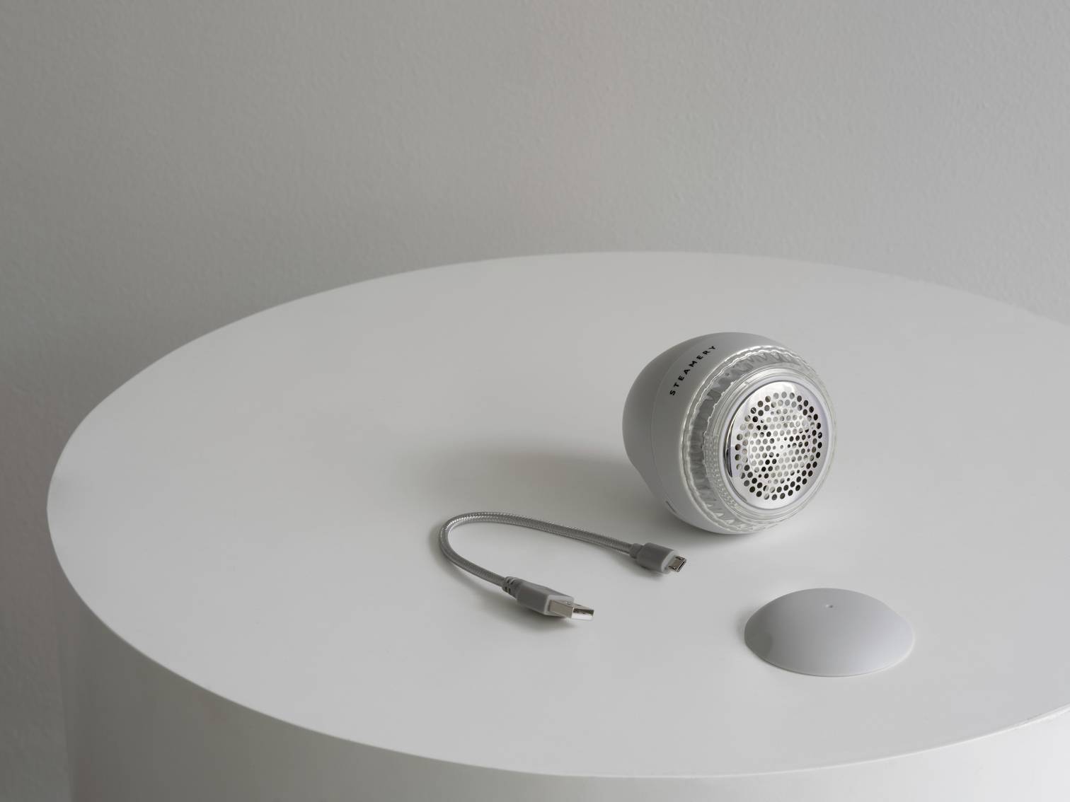 Grey, round speaker lying on a white table. Next to it, a USB cable and a detachable part of the speaker.