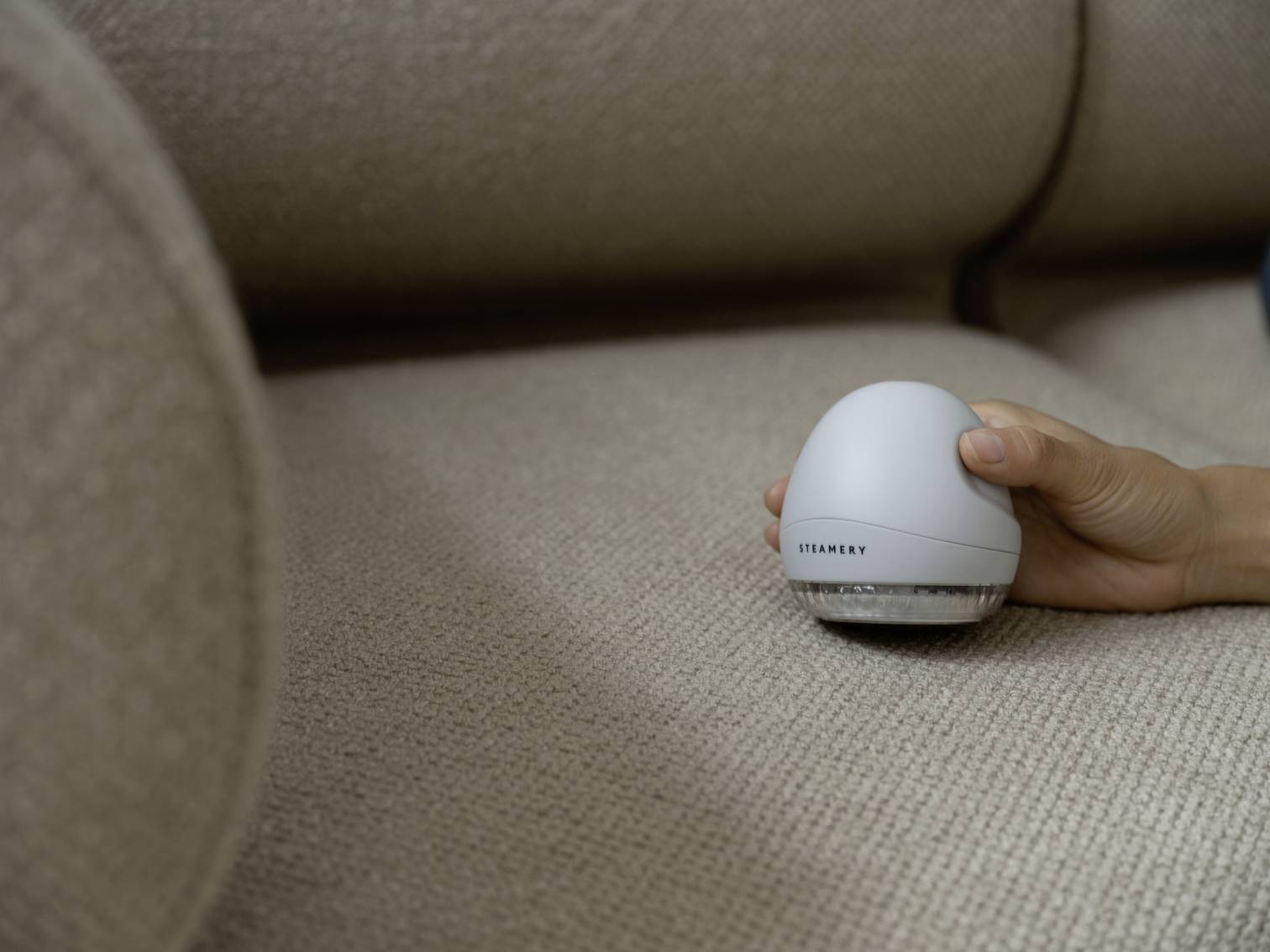A hand is holding a small, white, spherical device on a beige fabric sofa. The device is marked with the inscription 'STANLEY'.