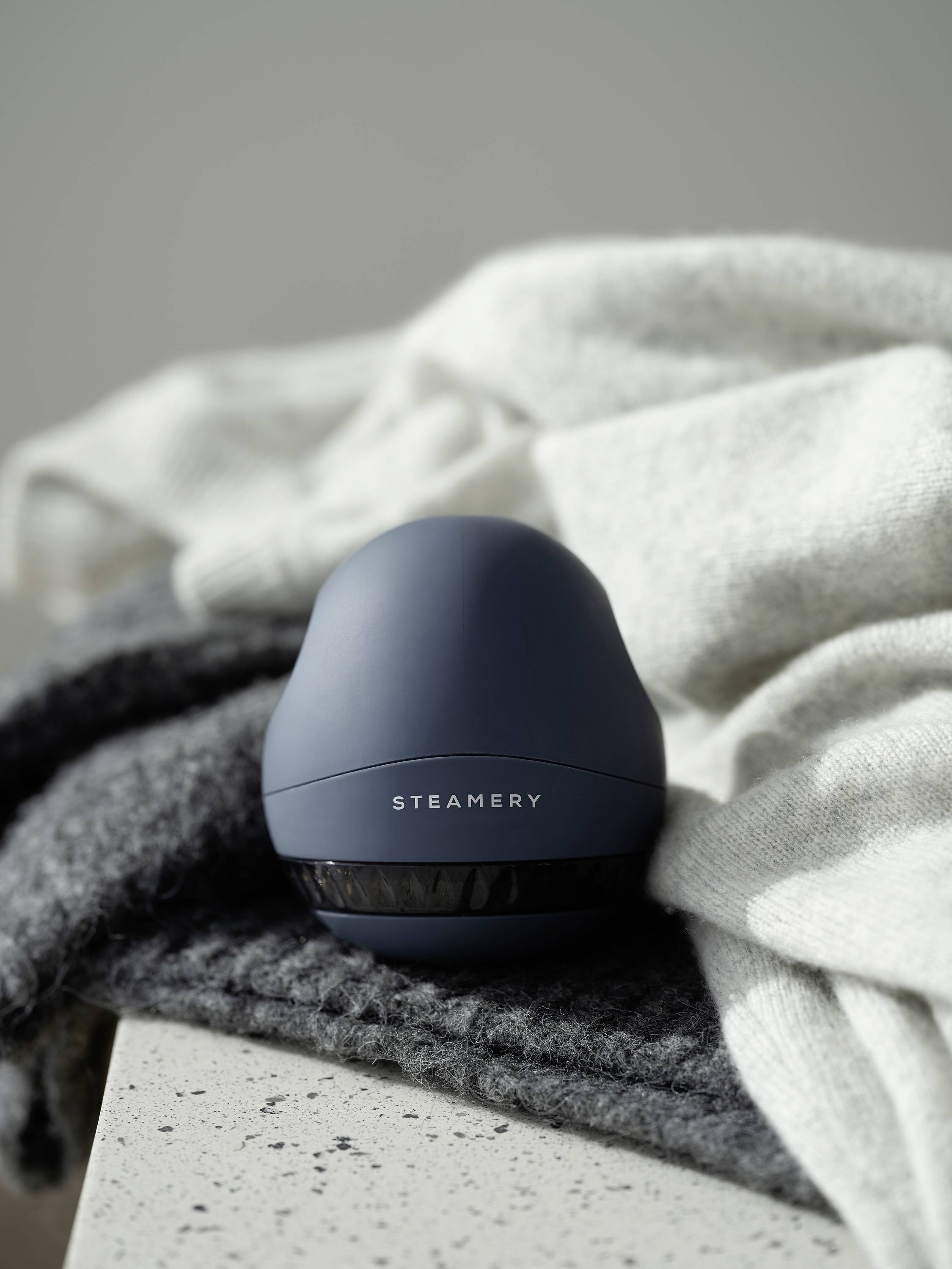 A blue lint remover on a grey fabric background. The device bears the inscription 'STEAMERY'.