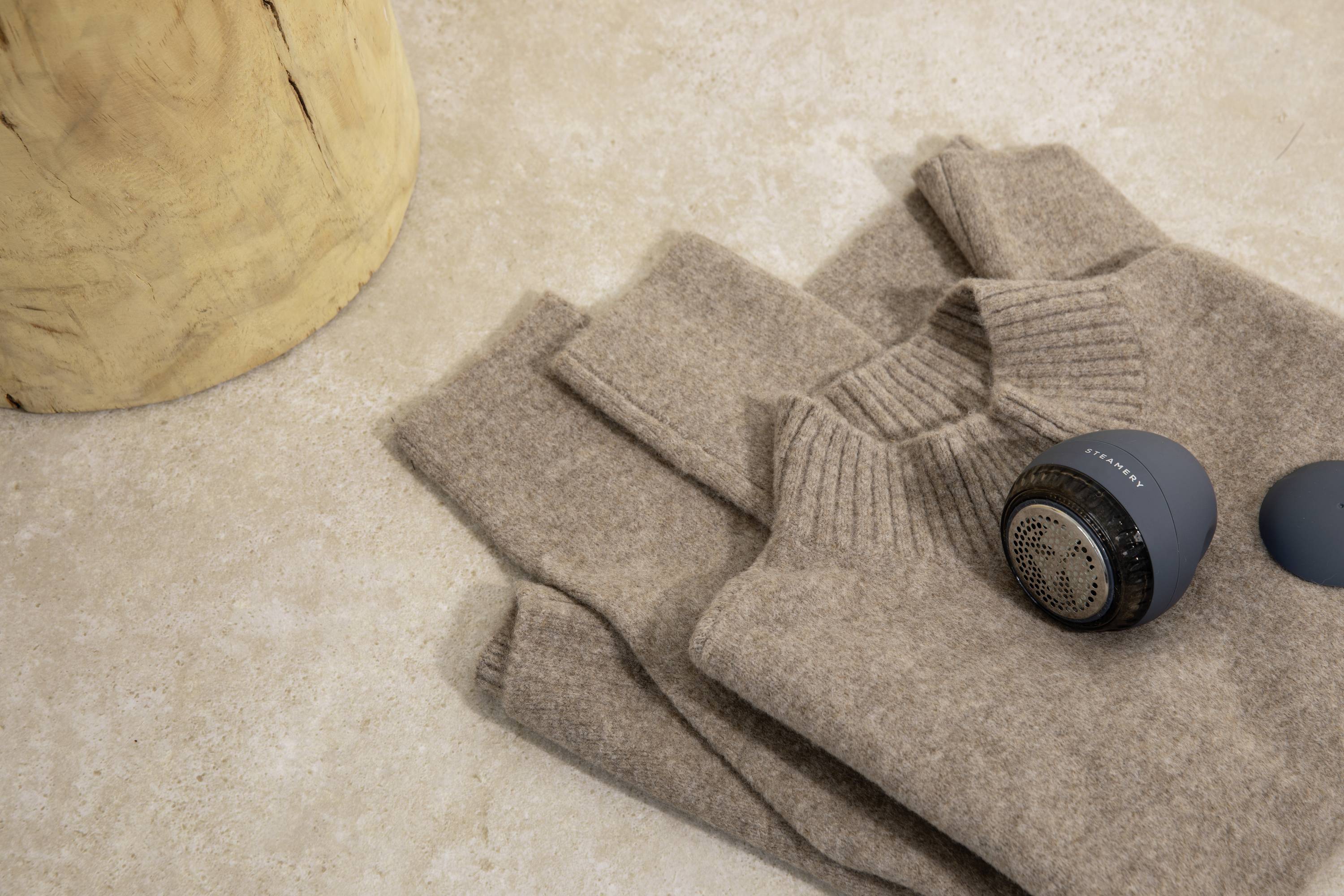 Beige jumper, folded next to a blue lint remover on a beige background, with a piece of wood visible on the left.