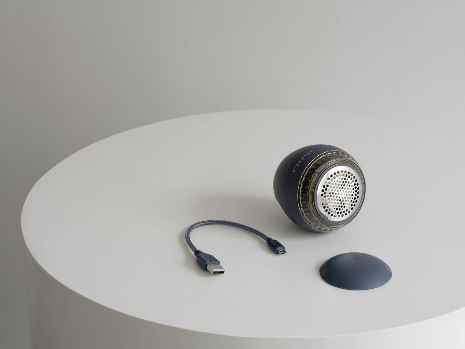 A small, black speaker is sitting on a white table. Next to it are a short USB cable and a removable cover.