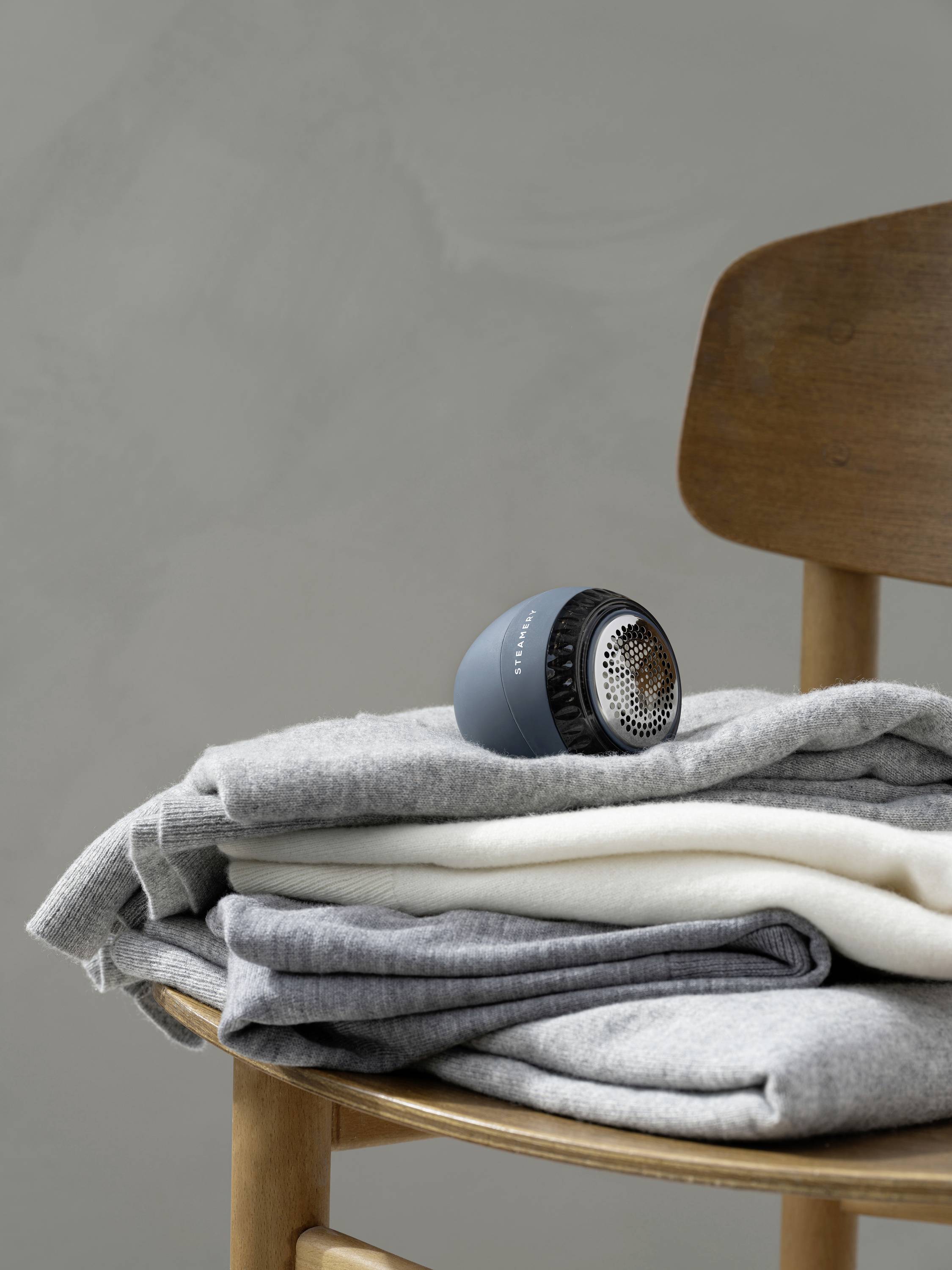 A wooden chair with a stack of folded grey and white clothing. On top sits a small, round electric lint shaver.
