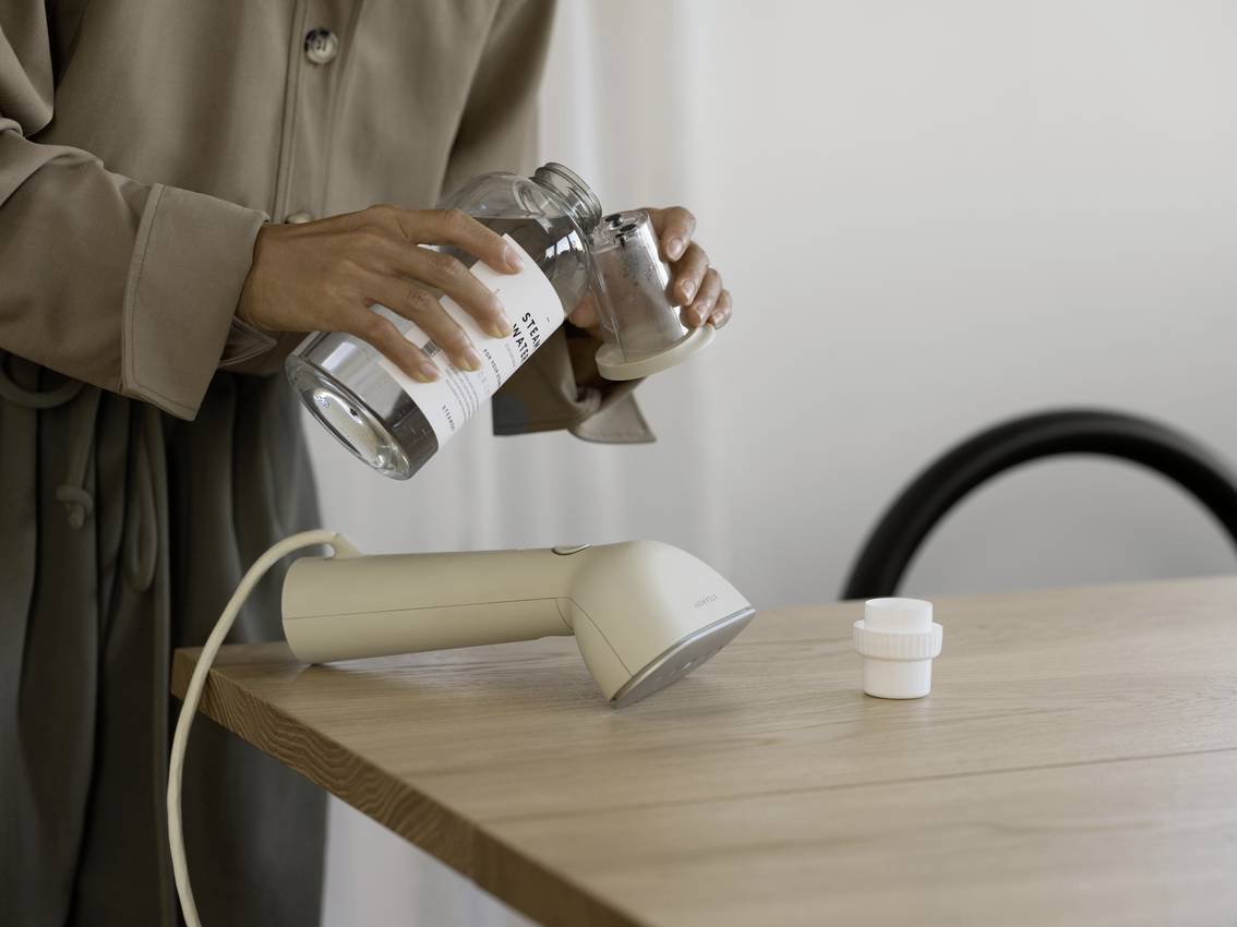 A person is pouring water from a bottle into a glass next to a steam iron on a wooden table. A plugged-in socket is visible.