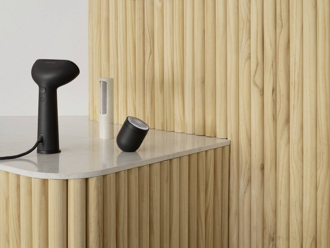 A black handheld steam cleaner and an accessory on a light wooden-panelled table with a wooden-clad background.