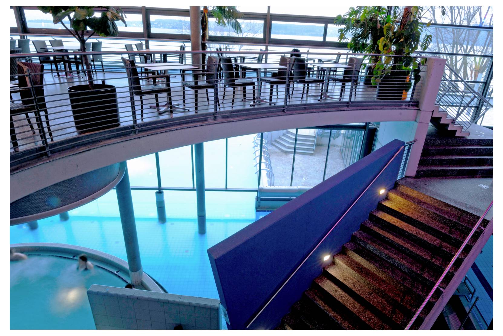 Interior of a modern building with a curved staircase beside a bright blue indoor pool or atrium, with a dining area above.