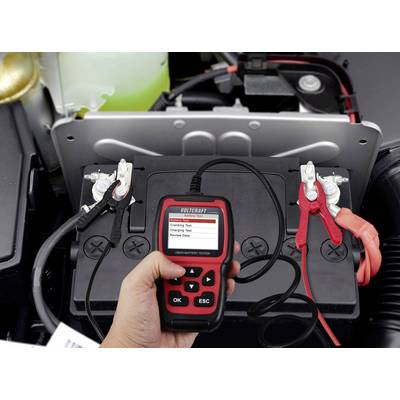 A hand is holding a battery testing device connected to a car battery. Various test options are displayed on the screen.