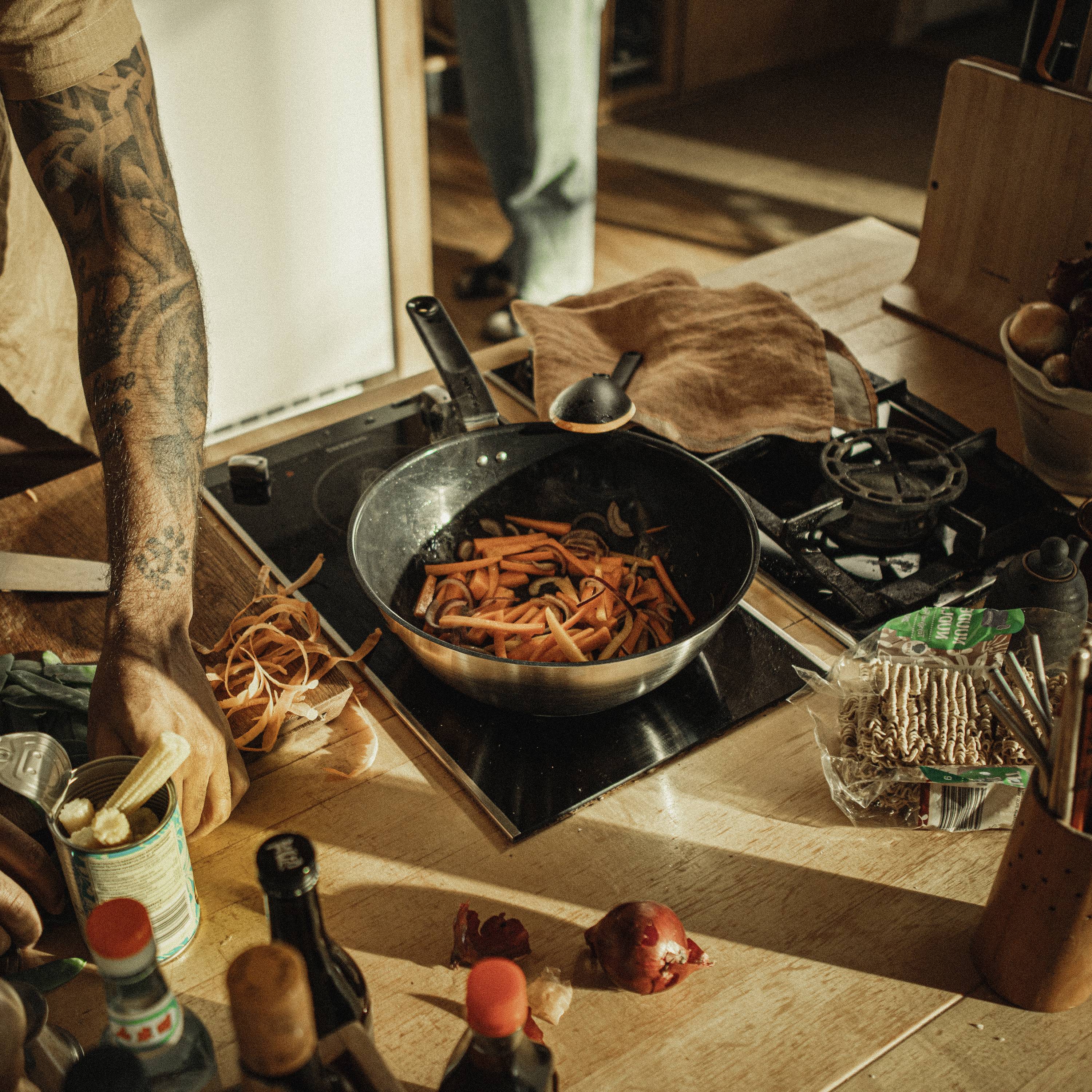A hand stirs carrot strips in a pan on a hob. Kitchen utensils and ingredients are scattered across a wooden worktop.