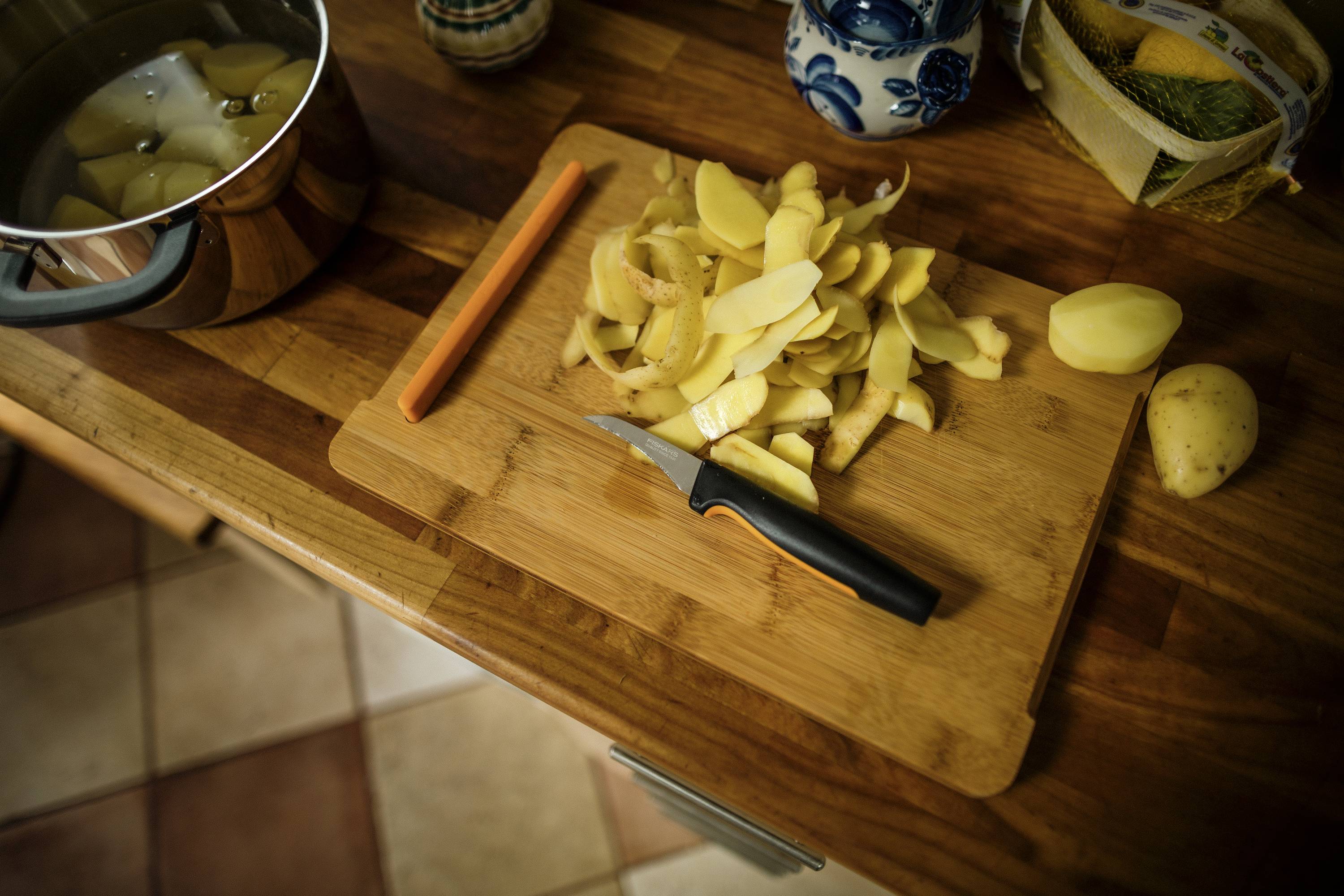 Potatoes are being boiled in a saucepan, alongside peeled potato skins on a wooden board and a small knife.
