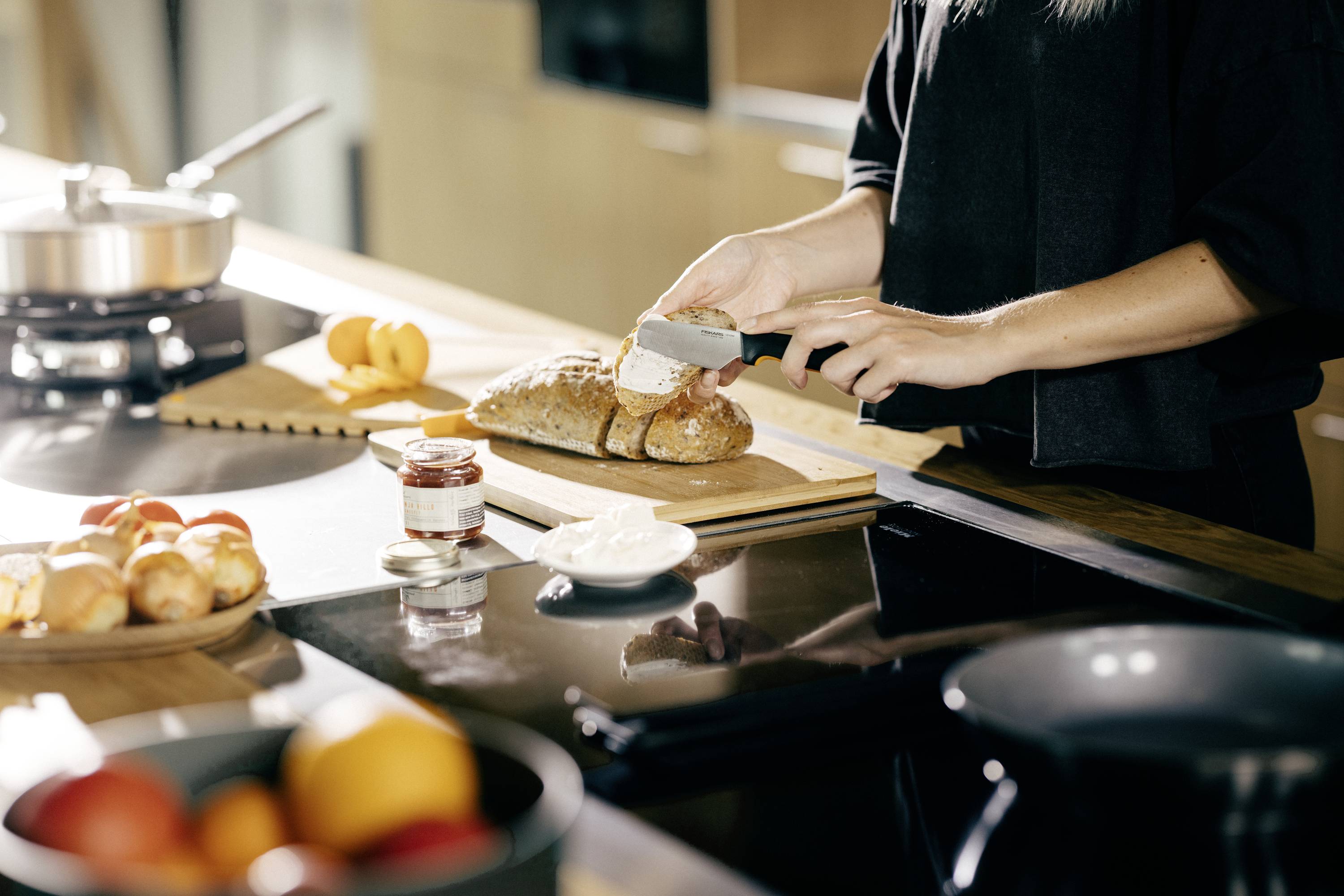 A person is cutting a fresh baguette in a kitchen. Jam and fruit are sitting on the table.