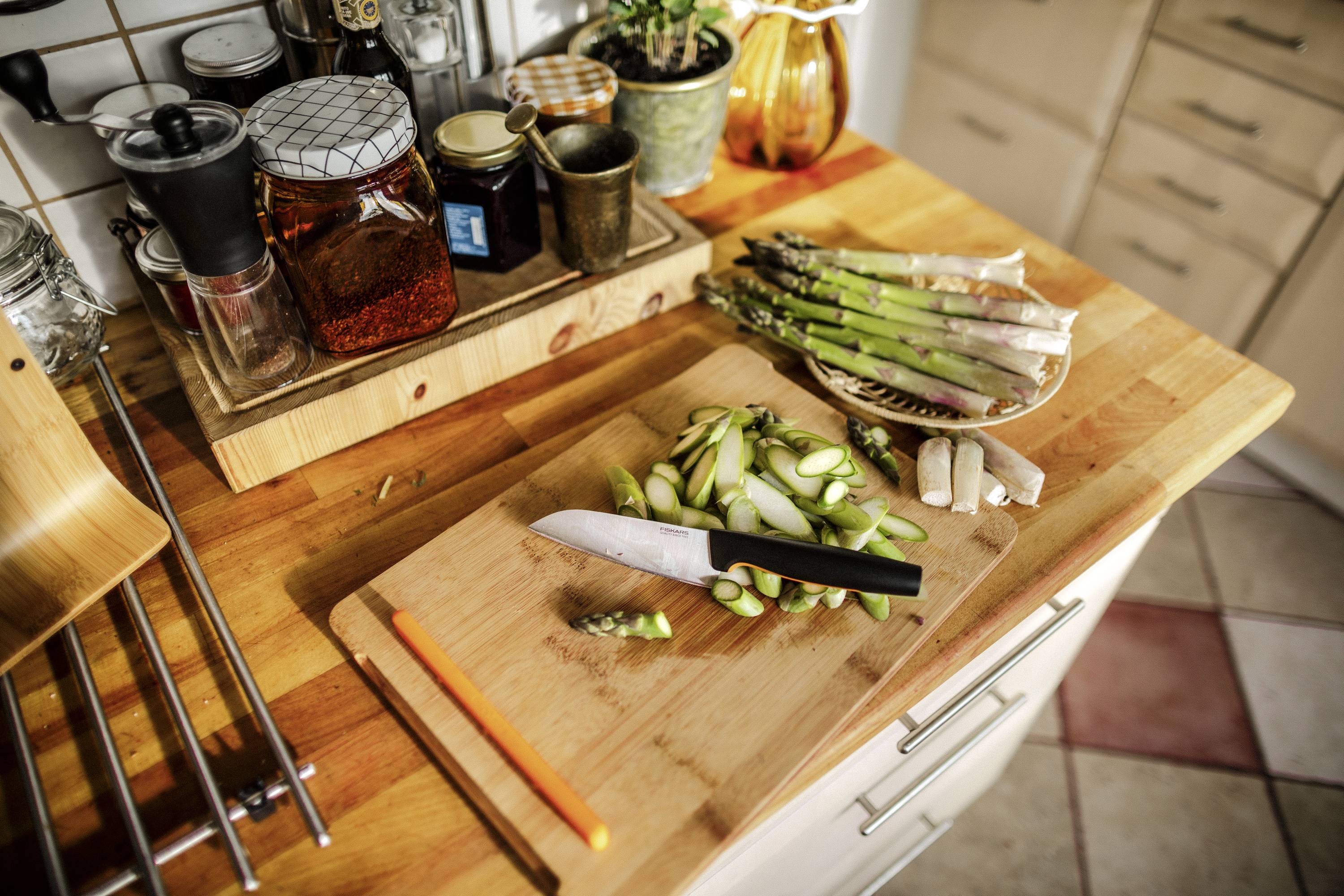 A kitchen table with sliced asparagus on a wooden chopping board, with a knife beside it. In the background, glasses of provisions are standing.