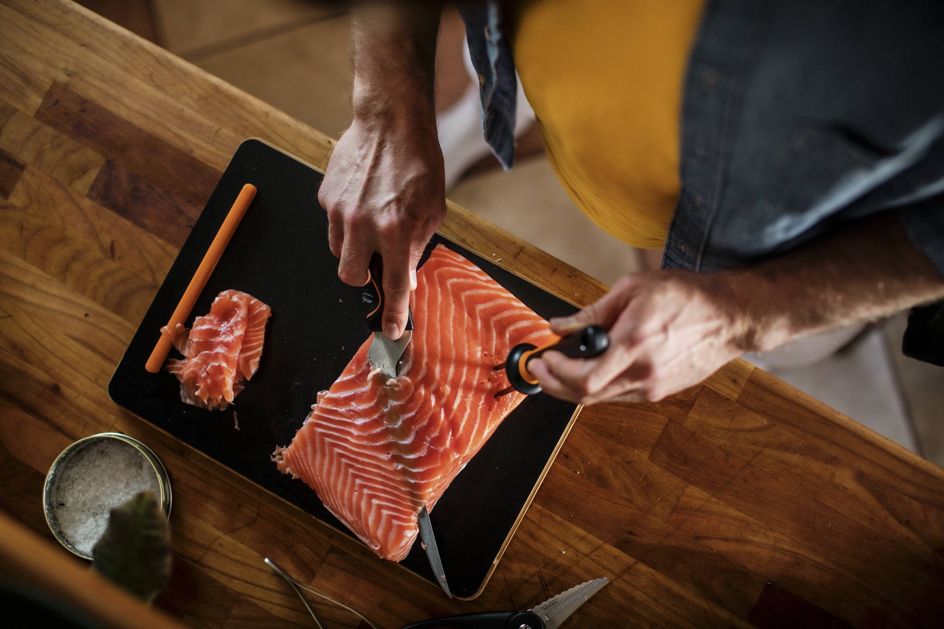A person is cutting salmon fillets on a chopping board. A smaller piece of salmon lies beside it, and an orange utensil is visible.