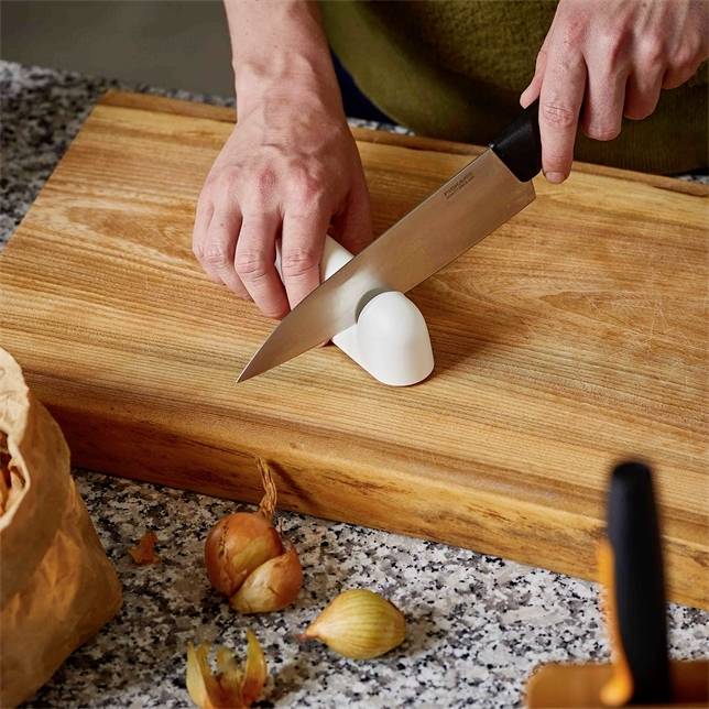 A person is chopping a white onion on a wooden chopping board with a knife. Some onions and a paper bag are in the foreground.