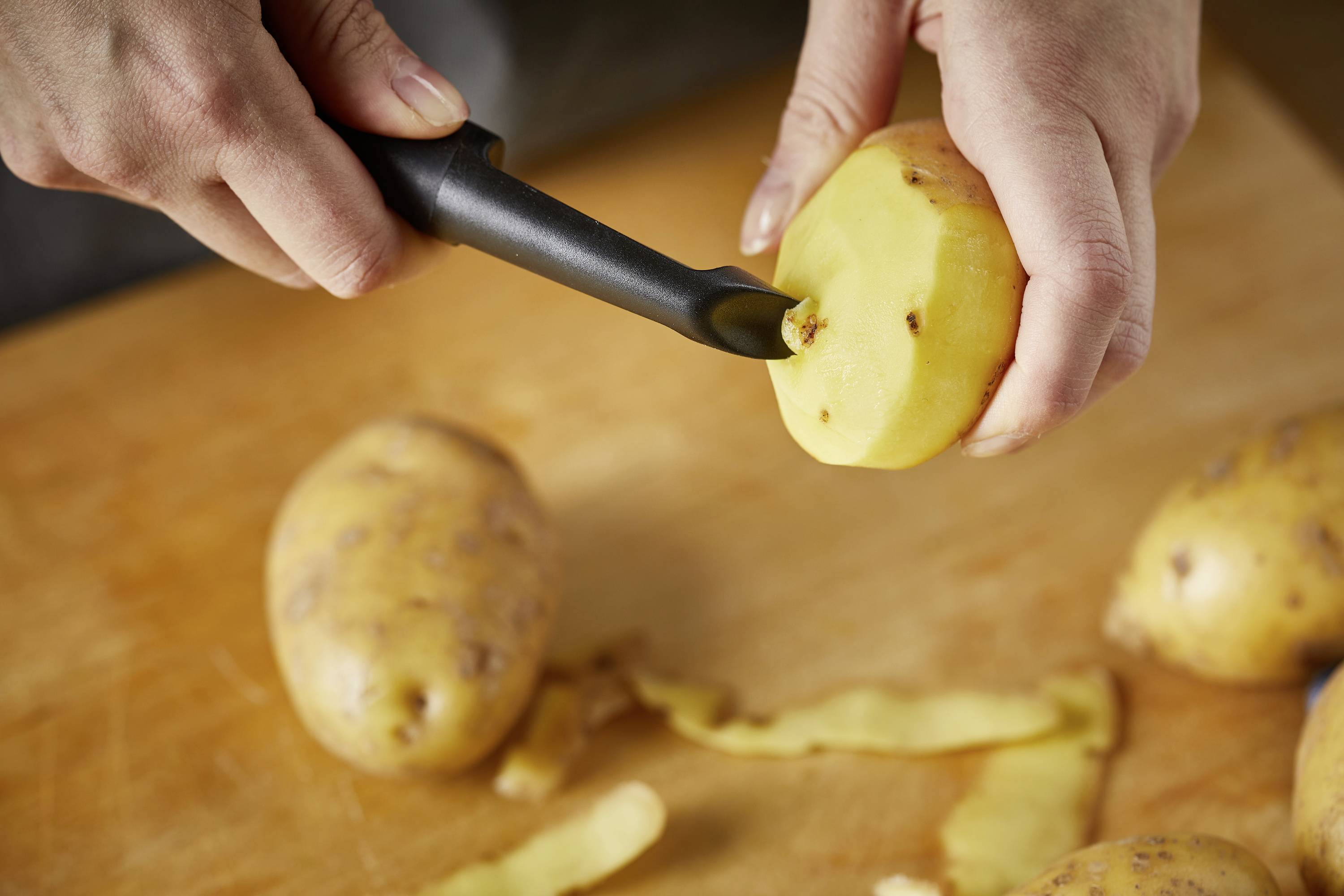 A person is peeling a potato with a vegetable peeler. Several unpeeled potatoes and potato peelings are visible on a wooden board.