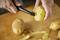 A person is peeling a potato with a vegetable peeler. Several unpeeled potatoes and potato peelings are visible on a wooden board.