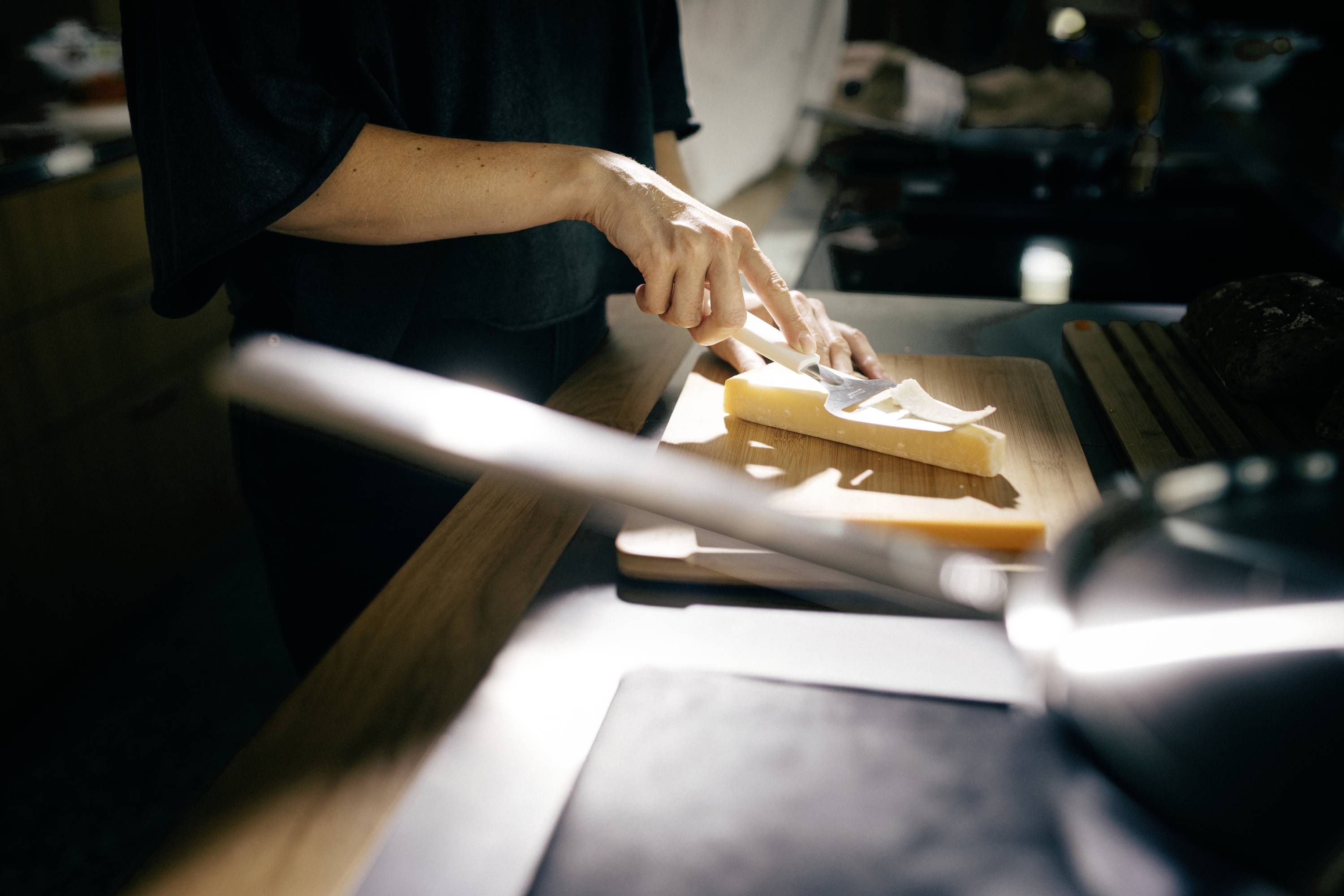 A person is cutting bread in a kitchen. Light falls from a window onto the chopping board. A frying pan is in the foreground.