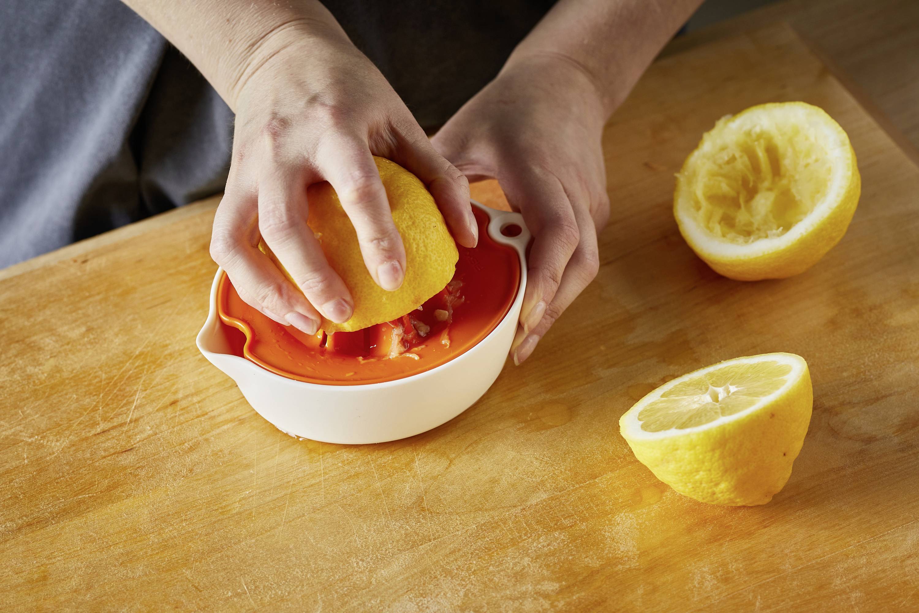 A person is squeezing half a lemon into a white juicer using both hands. Two other lemon halves are lying beside it on a wooden table.