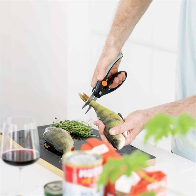 A person is cutting the fins off fish with kitchen scissors on a chopping board. A glass of red wine is in the foreground.