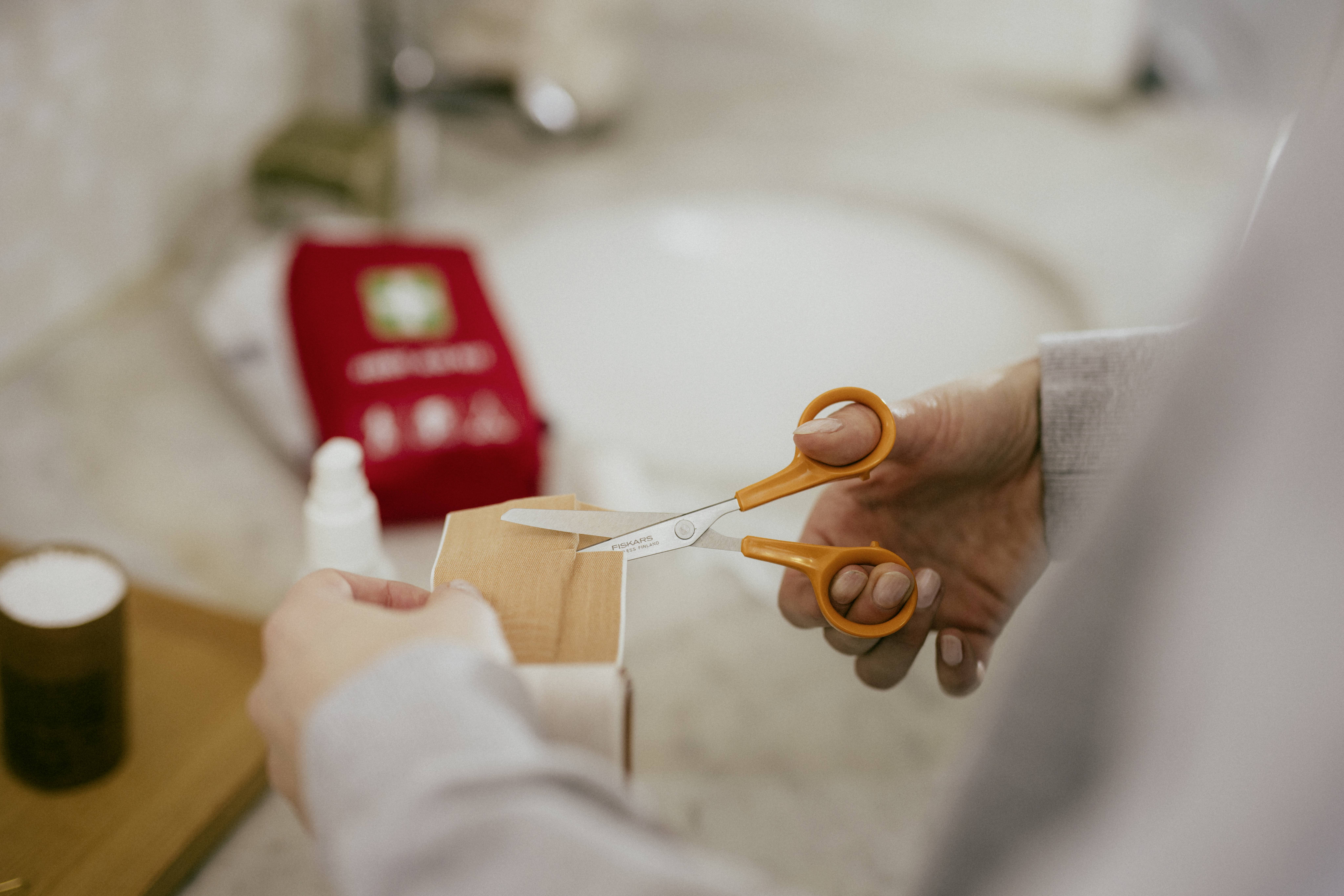A person is cutting a plaster (bandage) with small scissors. In the background, a red first aid kit is visible.