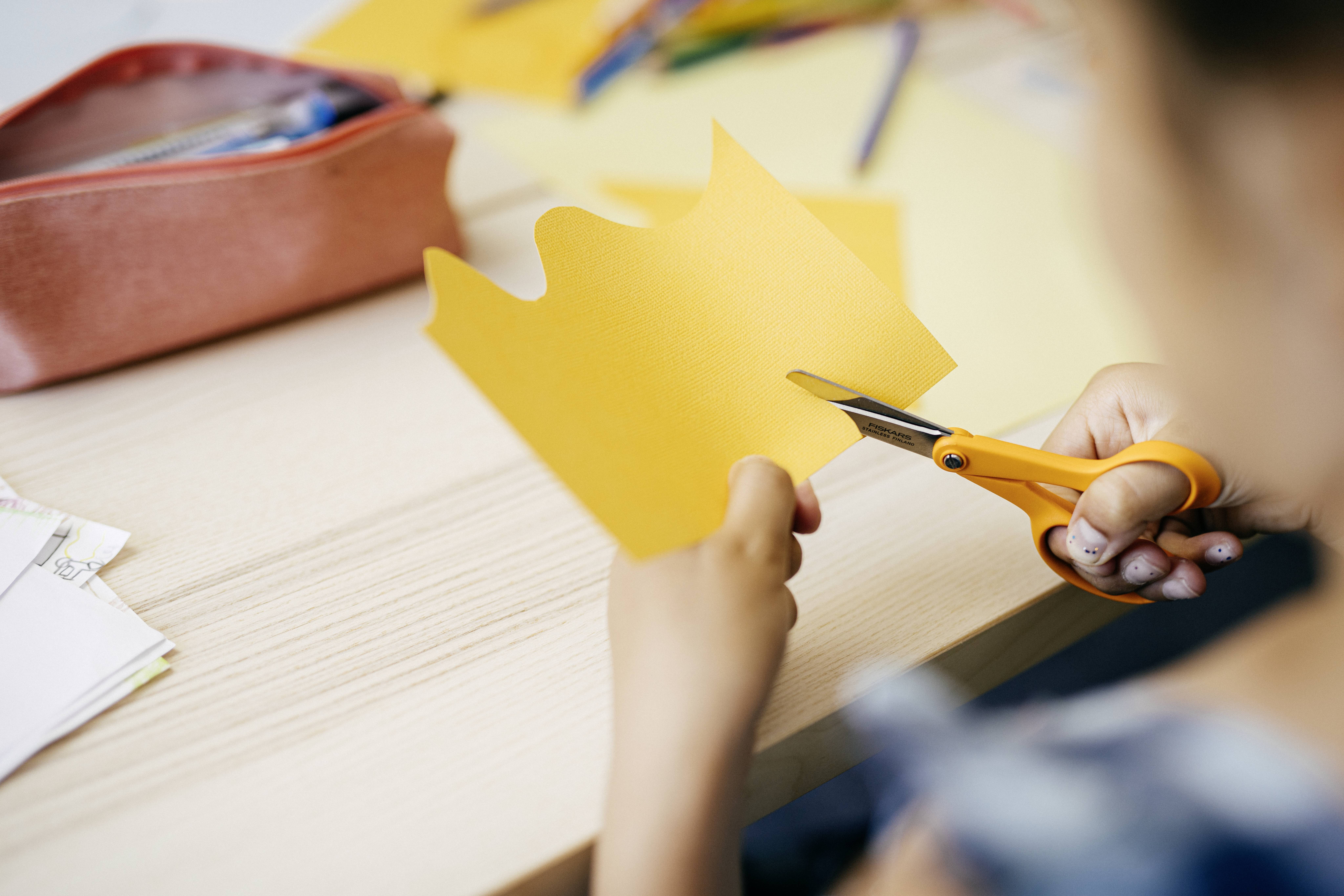 A child is cutting shapes out of yellow paper with scissors. In the background, a pencil case lies on the table.
