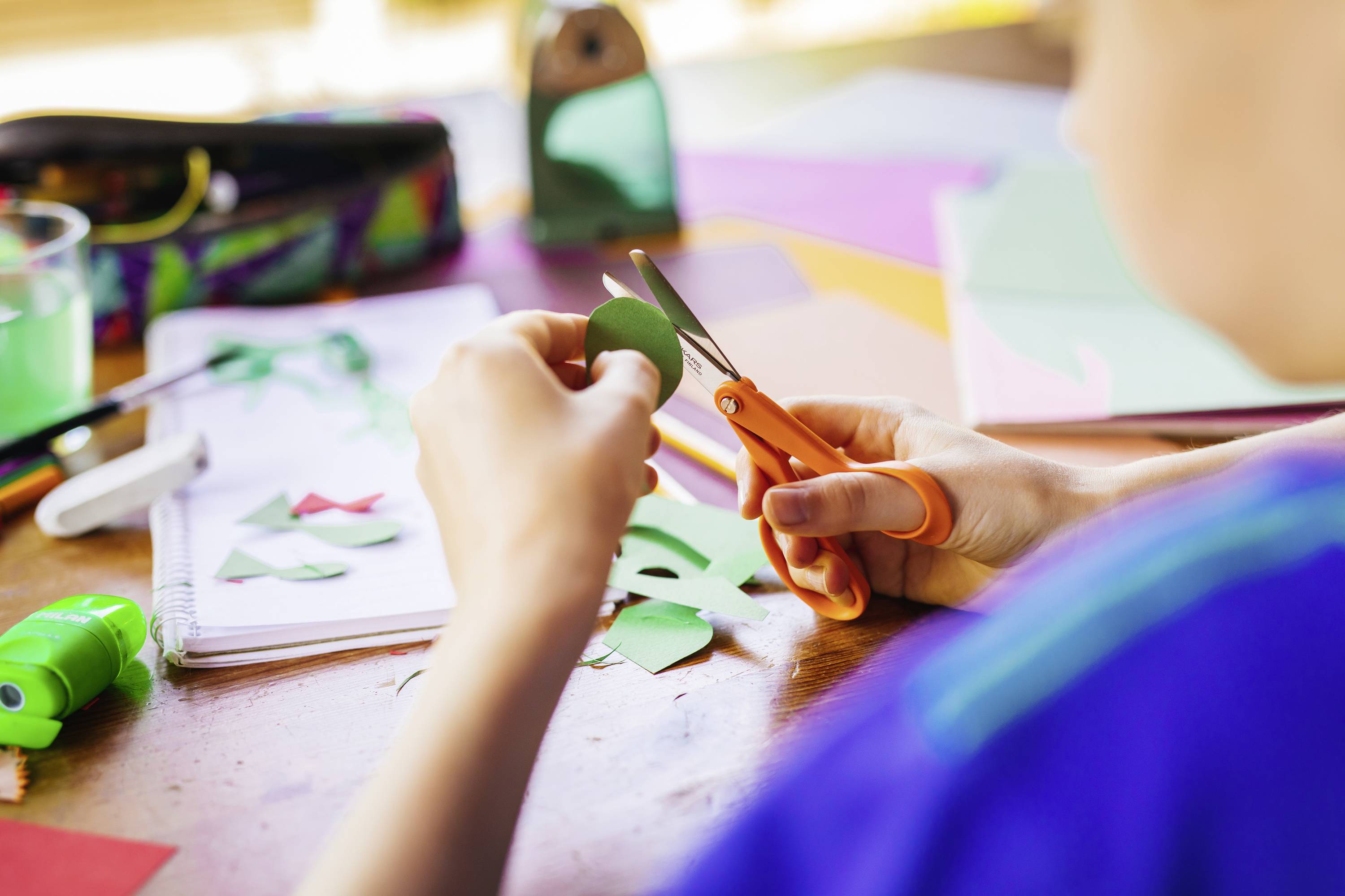 A person is cutting coloured paper with scissors. Various craft materials are spread out on a table in the background.