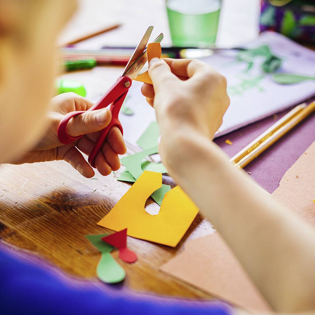 A person is cutting colourful paper with scissors. Additional craft materials and pens are lying on the table.