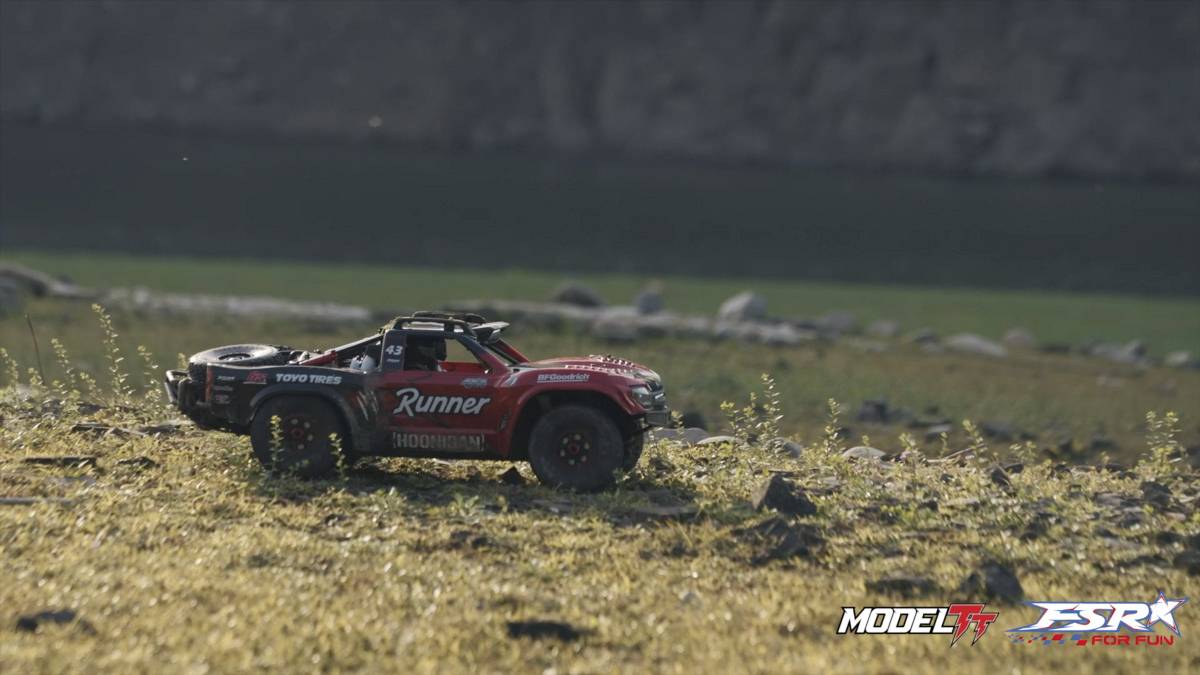 A red, remote-controlled car with the inscription 'Runner' drives across a meadow-like landscape with a blurred background.