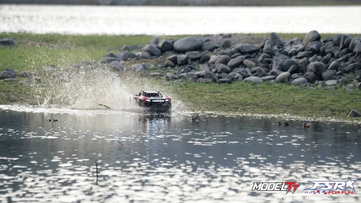 A remote-controlled car drives through shallow water, splashing water, surrounded by stones and a lake in the background.