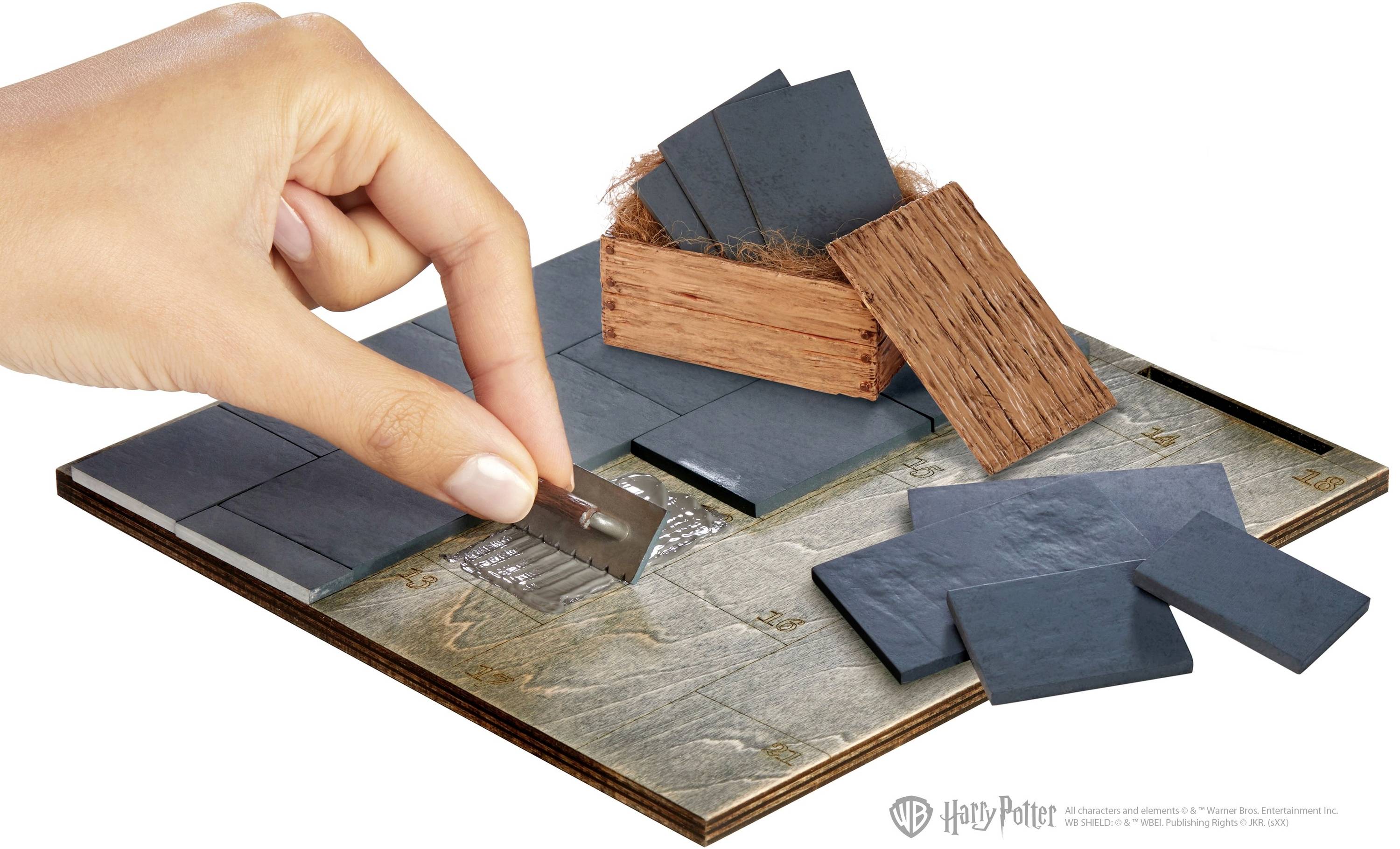 A hand holds a razor blade and cuts a rectangular, black, thin object from a tile on a wooden board.