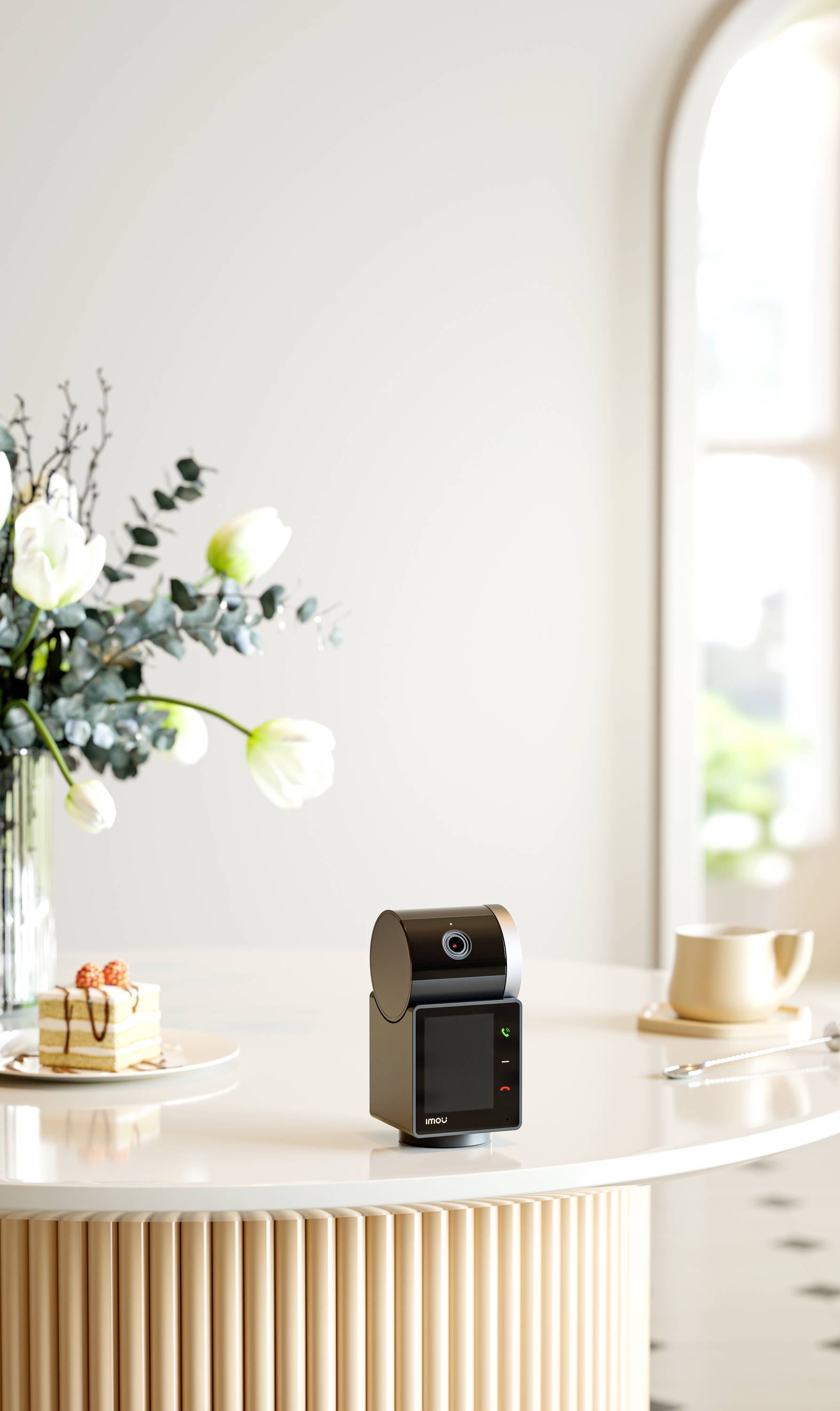 A small black speaker sits on a round table next to a cup of tea, a slice of cake, and a flower vase in front of a window.
