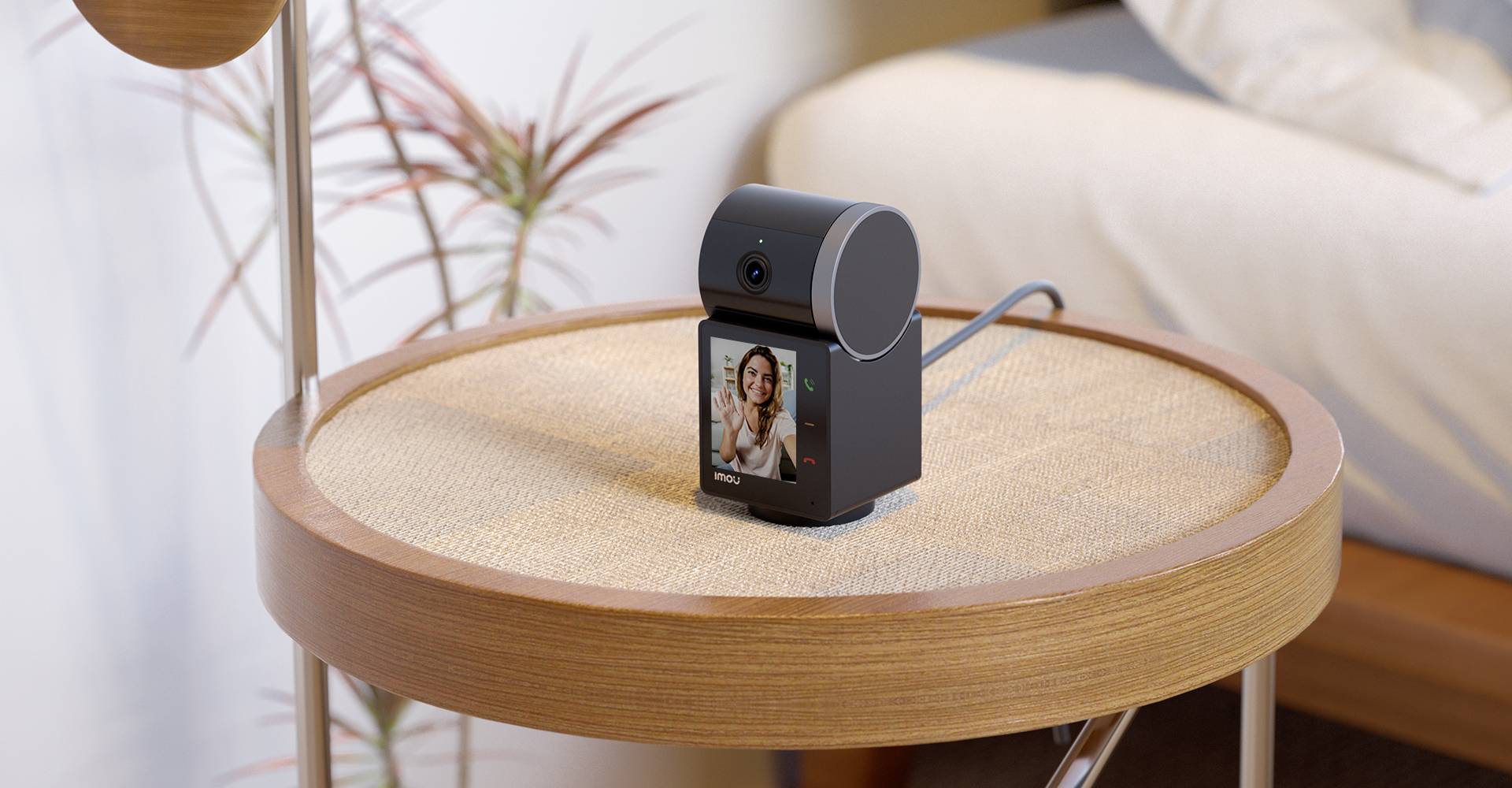A smart camera sits on a round wooden table. On its screen, the image of a person during a video call is visible.