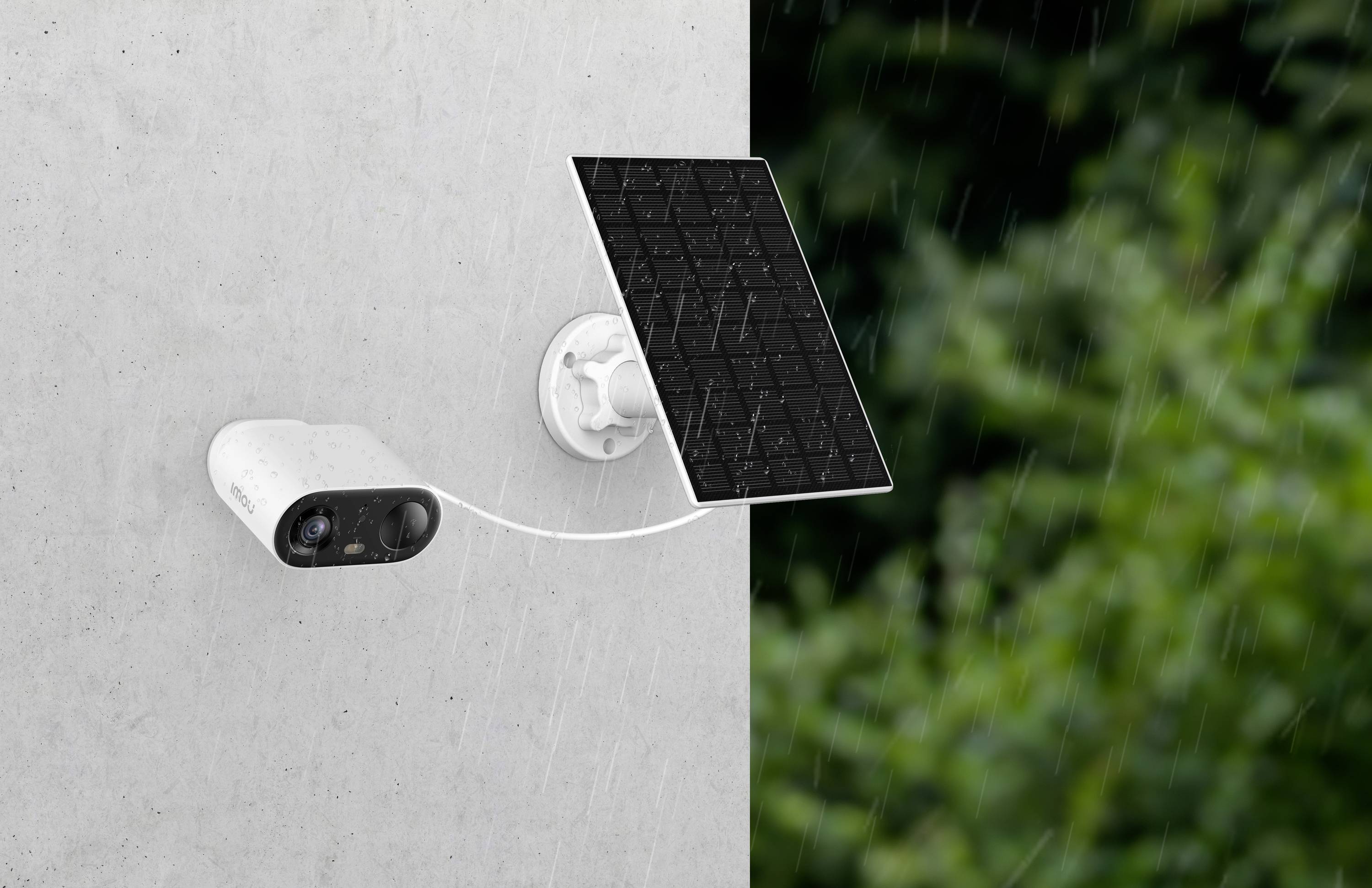 Security camera with solar panel mounted on a wall, in the rain in an outdoor area with green vegetation in the background.