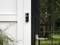 A smart home doorbell is installed on the exterior of a house in the rain, near a black glass door.
