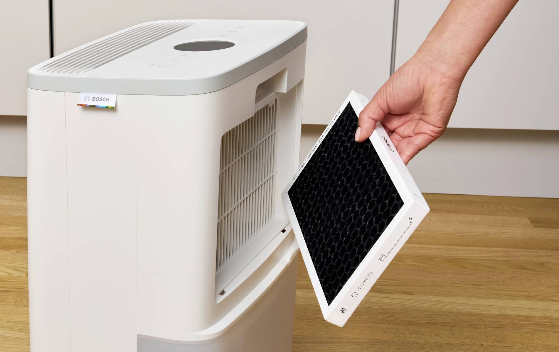 A person is changing the filter of an air purifier in a room with a wooden floor. The opened air purifier shows the removed filter.