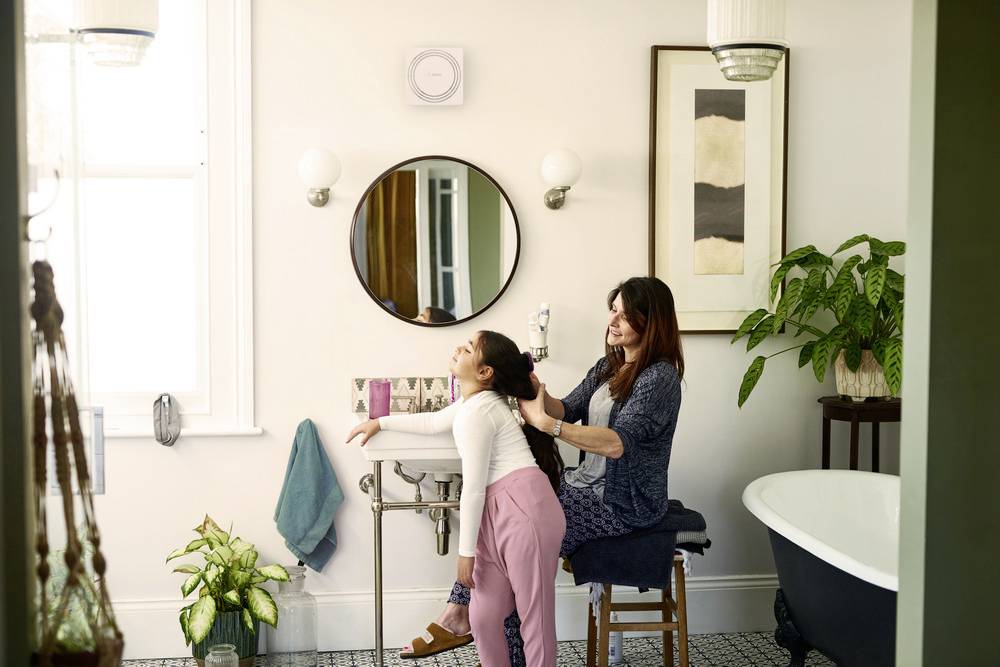 A woman is styling a child's hair in a bathroom with plants and a bathtub.