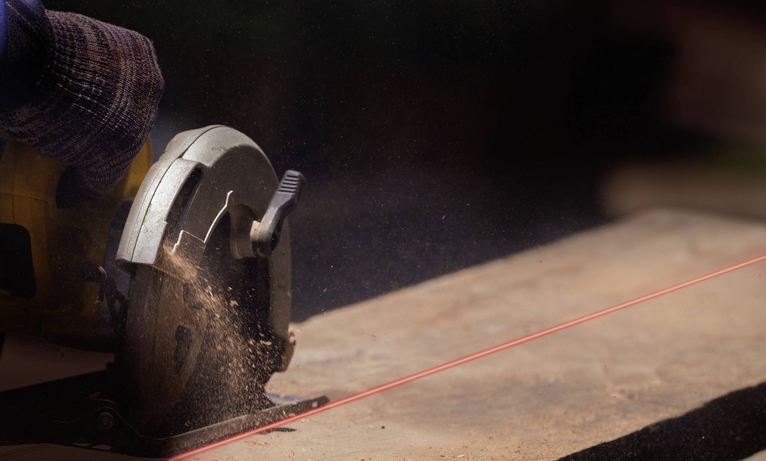 A circular saw cuts wood along a red laser line. Sawdust flies. Safety gloves protect the operator's hand.