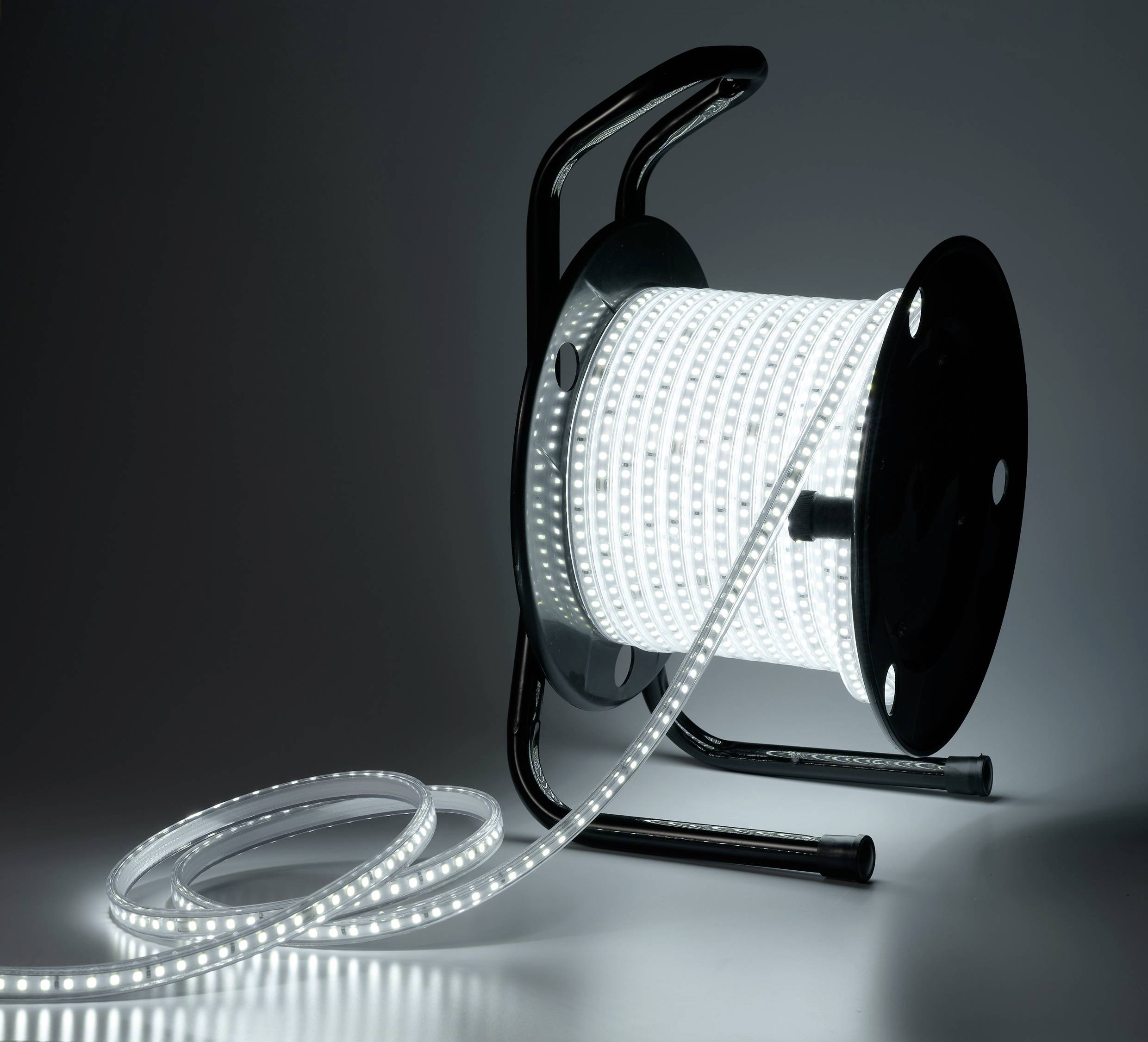 A luminous LED strip is wound around a black cable reel, unspooled on the floor. The background is dark.