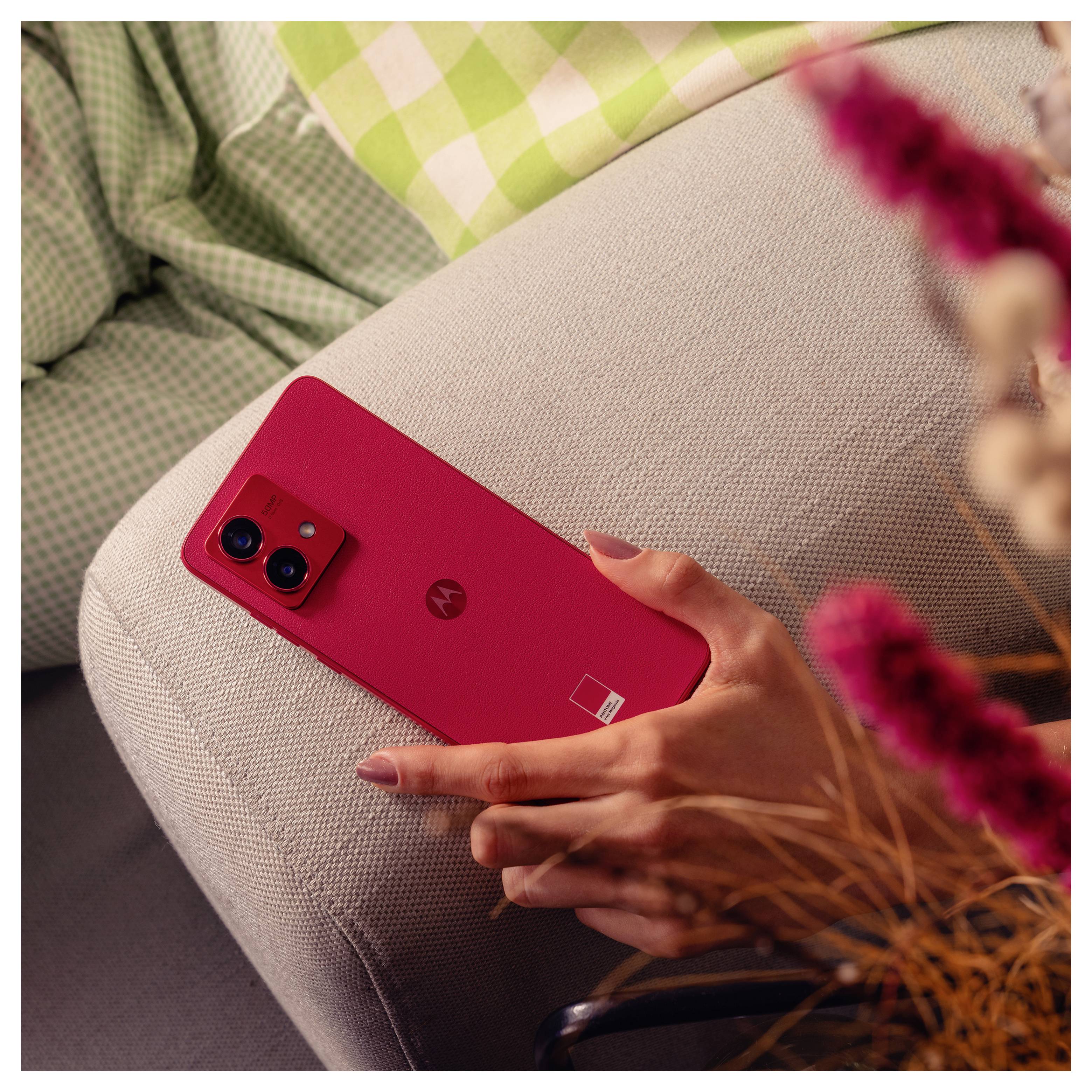 A person holds a red smartphone with a dual camera on a light-colored sofa. Nearby are green and white checkered fabric and dried flowers.