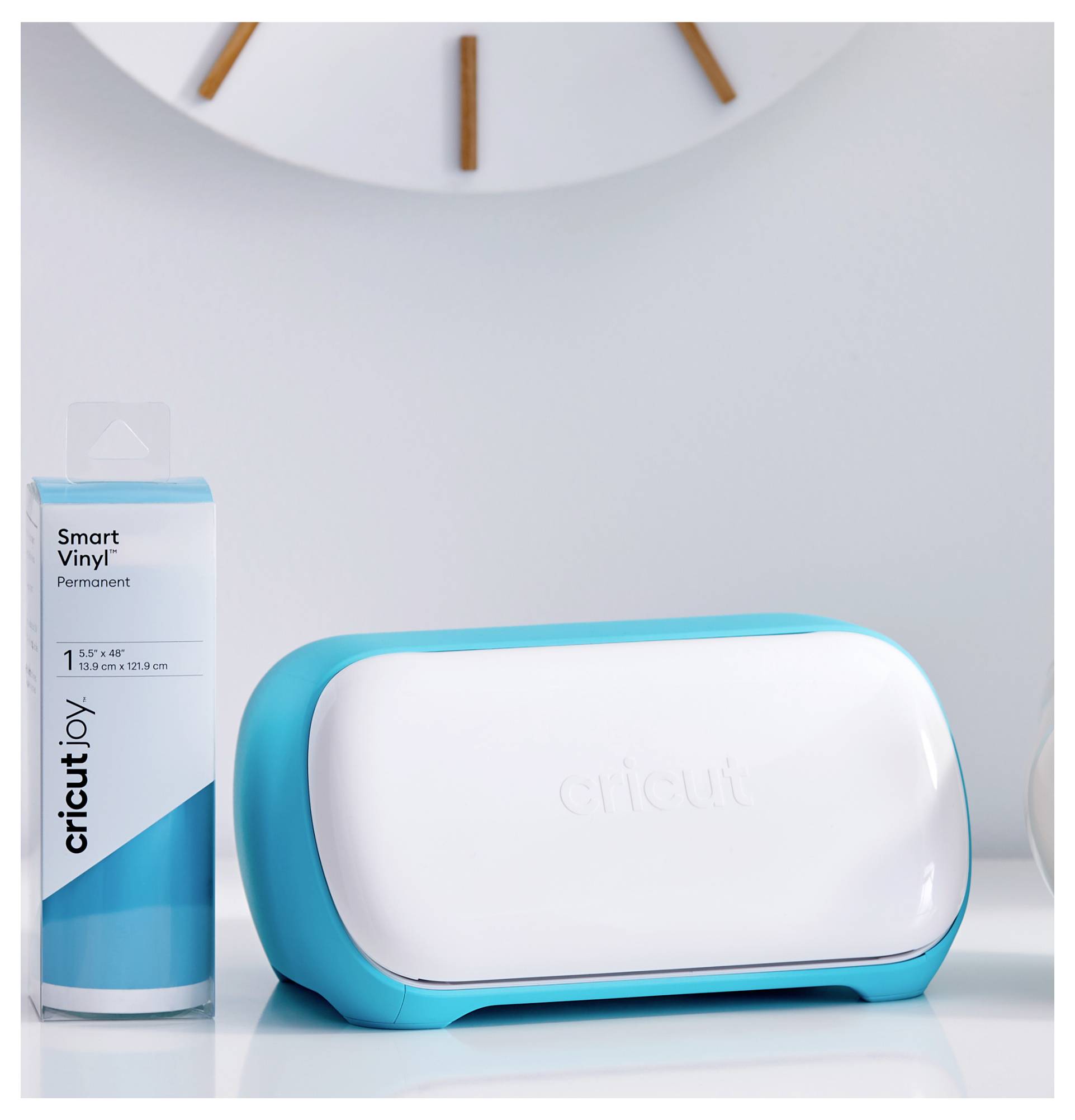 A 'Cricut Joy' cutting machine in blue and white sits on a table next to a pack of Smart Vinyl, with a clock in the background.